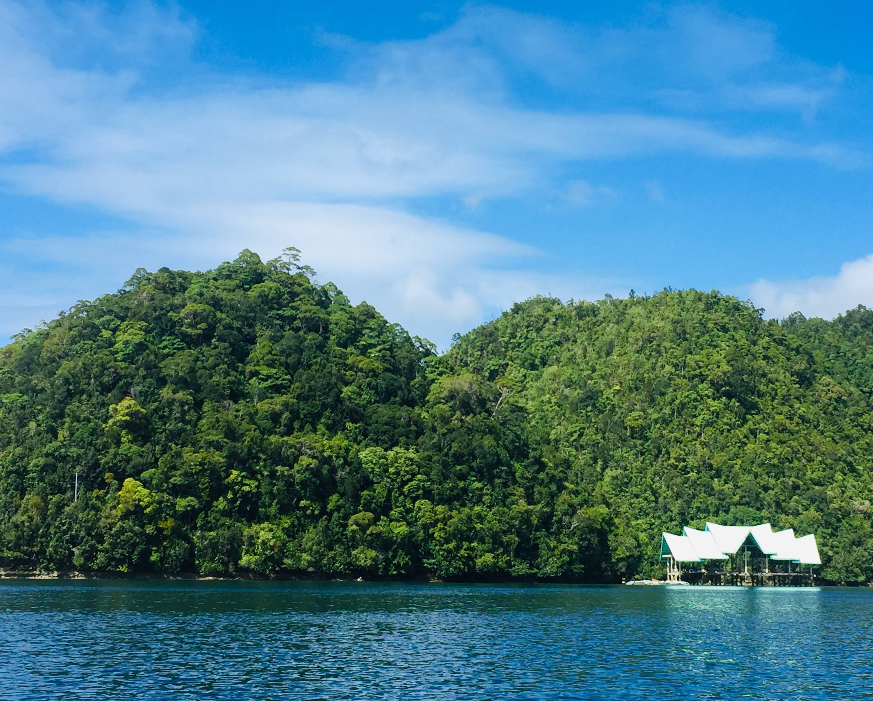Siargao 桌布 壁紙 — 一個寧靜的熱帶湖泊，周圍環繞著鬱鬱蔥蔥的綠色山丘，一座白色屋頂的高腳屋漂浮在平靜的藍色水面上，天空湛藍。