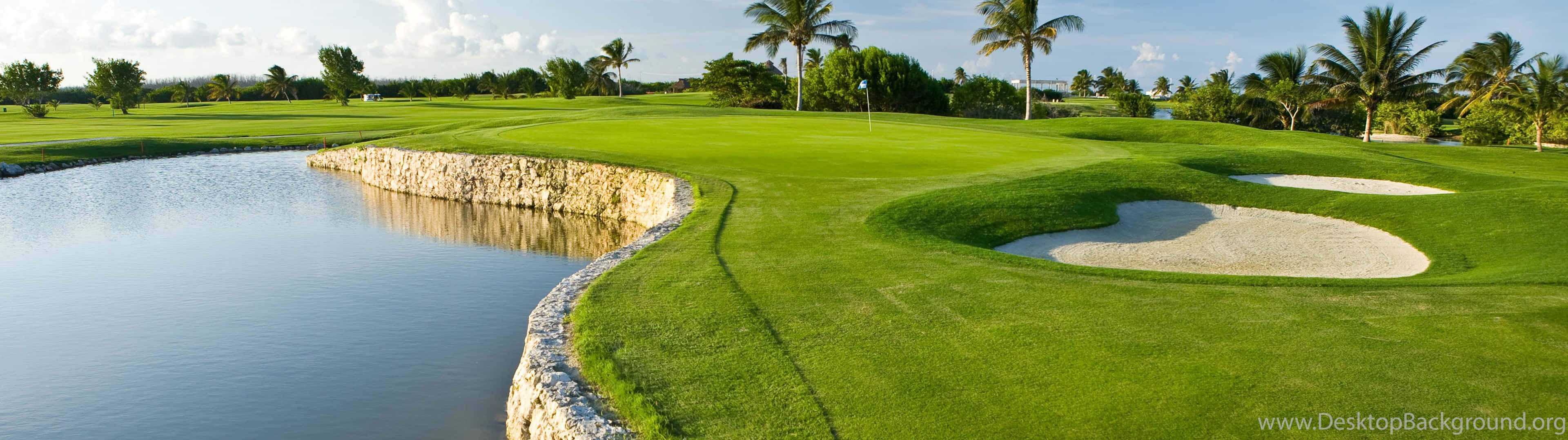 Golf 3840x1080 桌布 壁紙 — 一個寧靜的熱帶高爾夫球場，擁有茂密的綠色球道、平靜的水障礙和棕櫚樹，在明亮的藍天下。