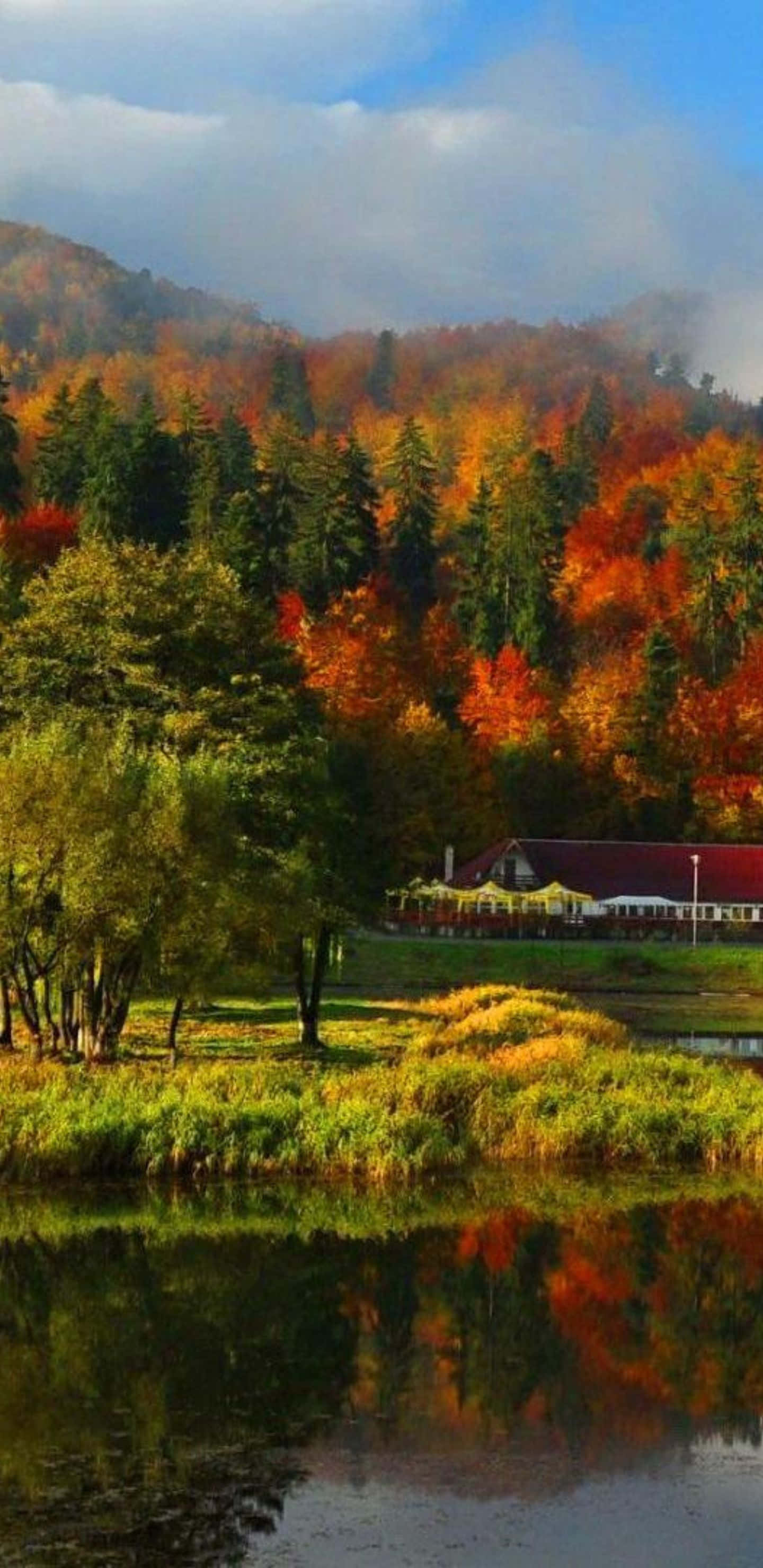 Beautiful Nature 桌布 壁紙 — 一個寧靜的秋日湖面倒映著環繞質樸小屋的火焰般紅色、橙色和綠色樹木，天空多雲。
