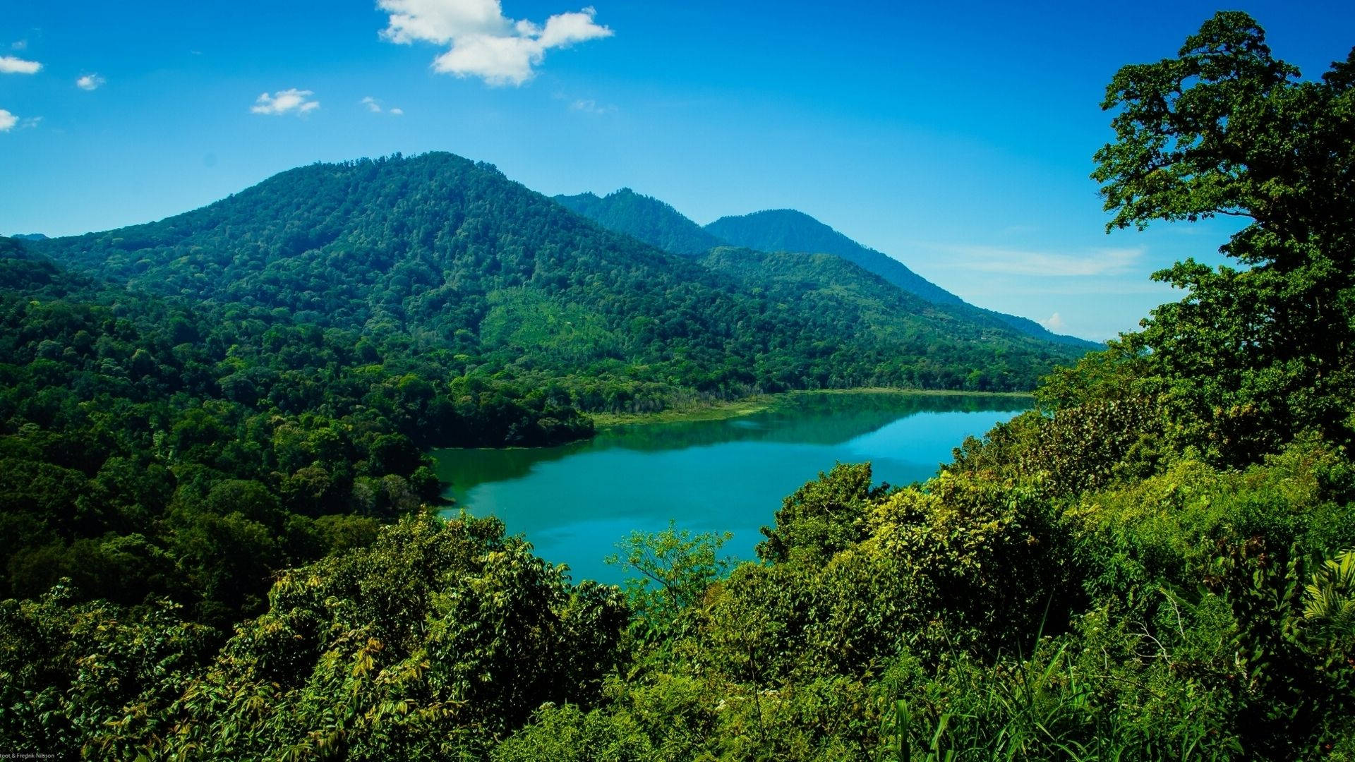 Jakarta 桌布 壁紙 — 一個寧靜的藍綠色湖泊，坐落在鬱鬱蔥蔥的綠色山脈之間，上方是明亮的藍天，點綴著幾朵雲彩。