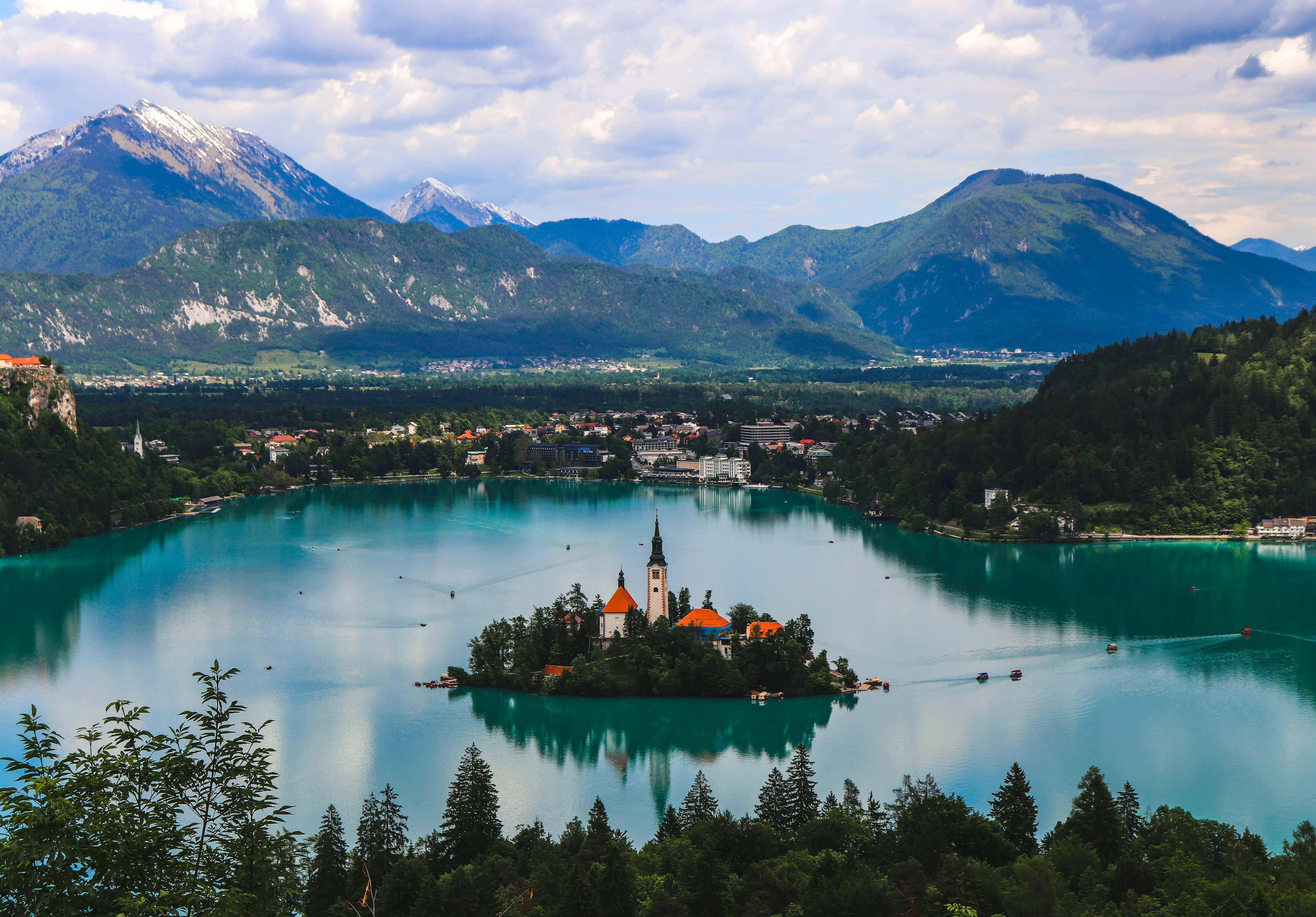 Slovenia 桌布 壁紙 — 一個寧靜的藍綠色湖泊懷抱著一個小島，島上有教堂，周圍是白雪覆蓋的山脈和茂密的綠色森林，天空部分多雲。