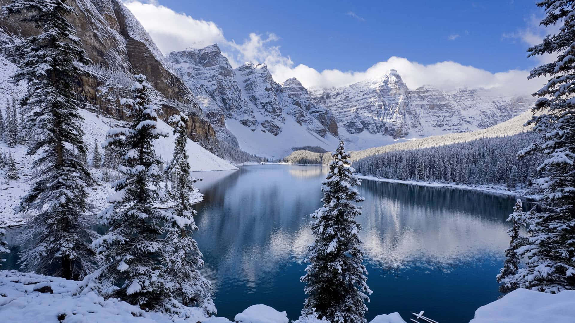 Best Winter 桌布 壁紙 — 一個寧靜的雪山湖泊倒映著高聳的山峰和常綠樹木，在明亮的藍天和蓬鬆的白雲下。
