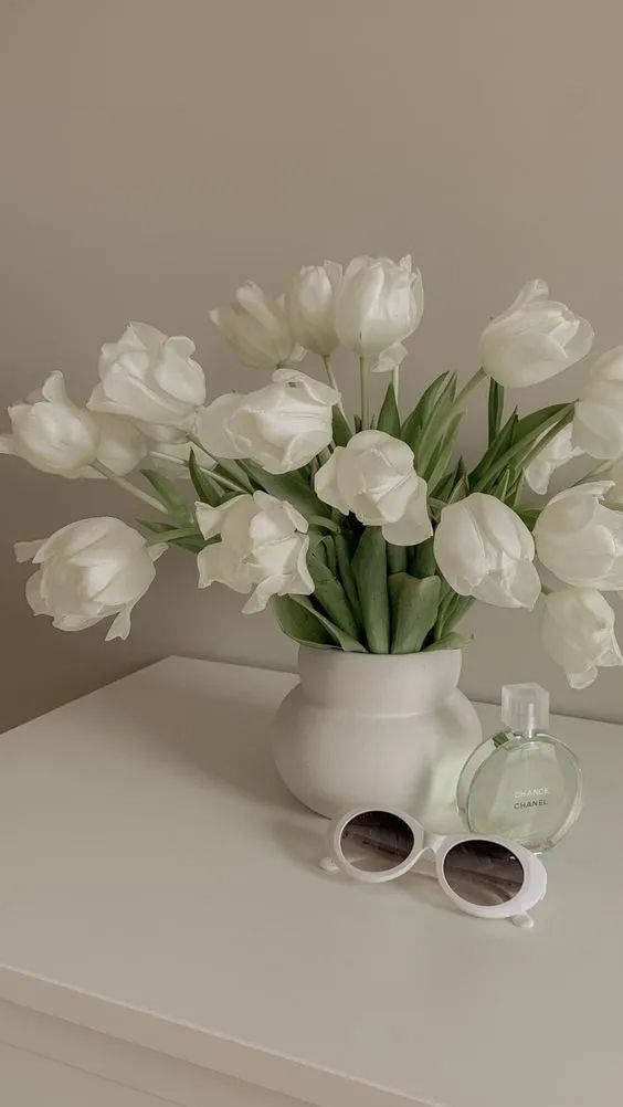 Flowers Aesthetic 桌布 壁紙 — 一個寧靜的靜物畫，白色鬱金香放在白色花瓶裡，旁邊有太陽眼鏡和香水瓶，放在簡約的白色架子上。