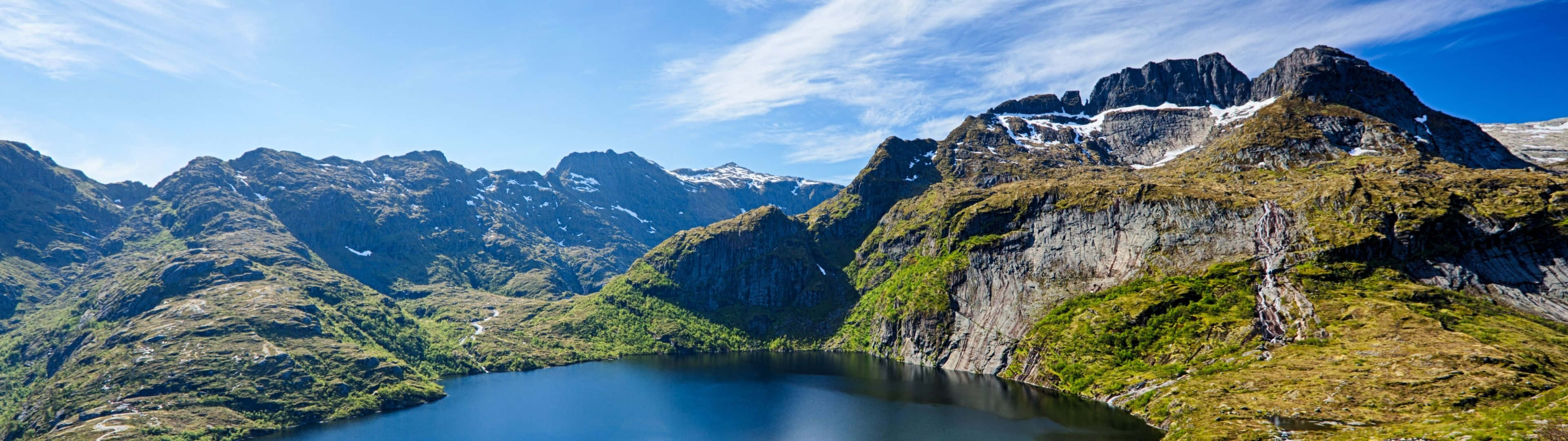 3840X1080 桌布 壁紙 — 一個寧靜的高山湖泊，在崎嶇的綠色和積雪覆蓋的山脈之間，映照出清澈的藍天。