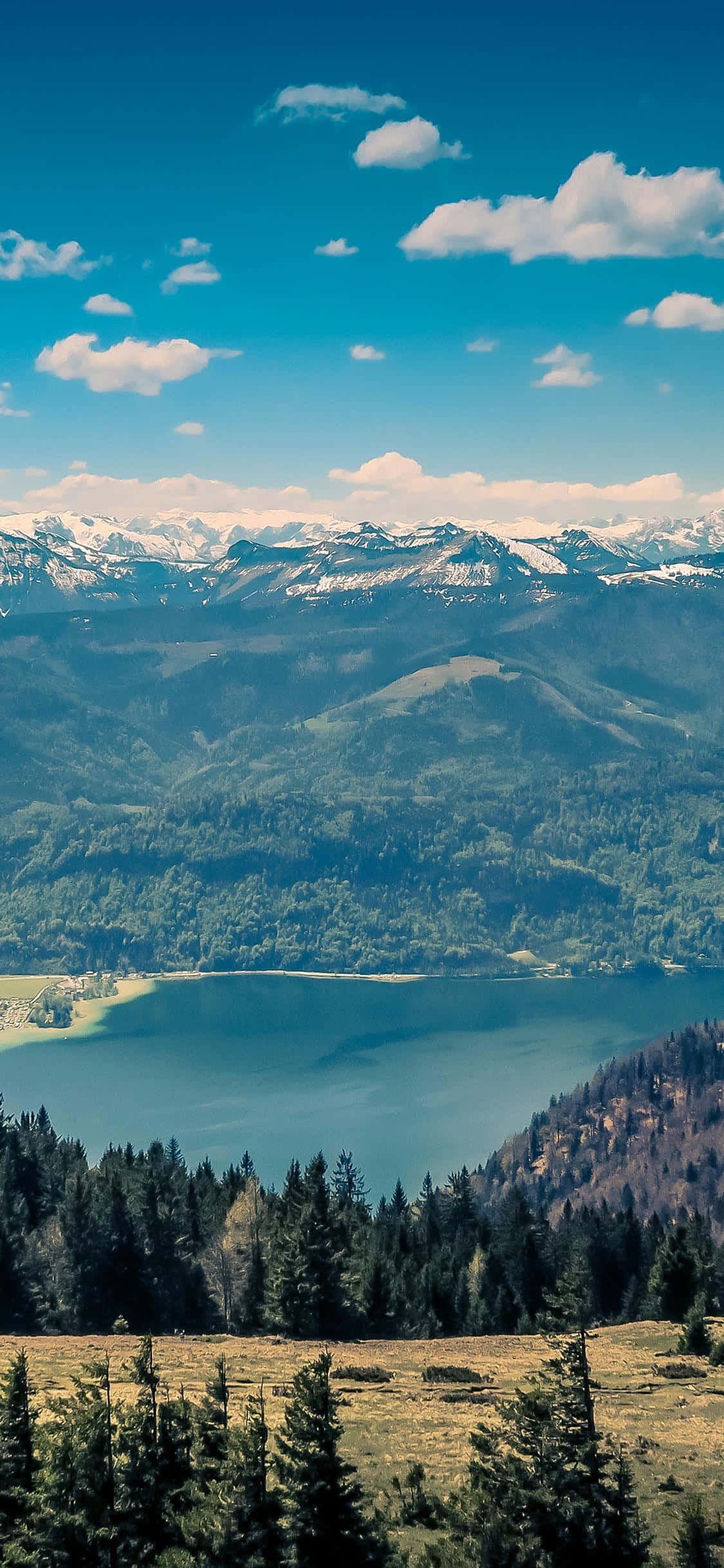 iPhone X Nature 桌布 壁紙 — 一個寧靜的高山湖泊，坐落在森林覆蓋的山丘和積雪覆蓋的山脈之間，天空湛藍，點綴著蓬鬆的白雲。