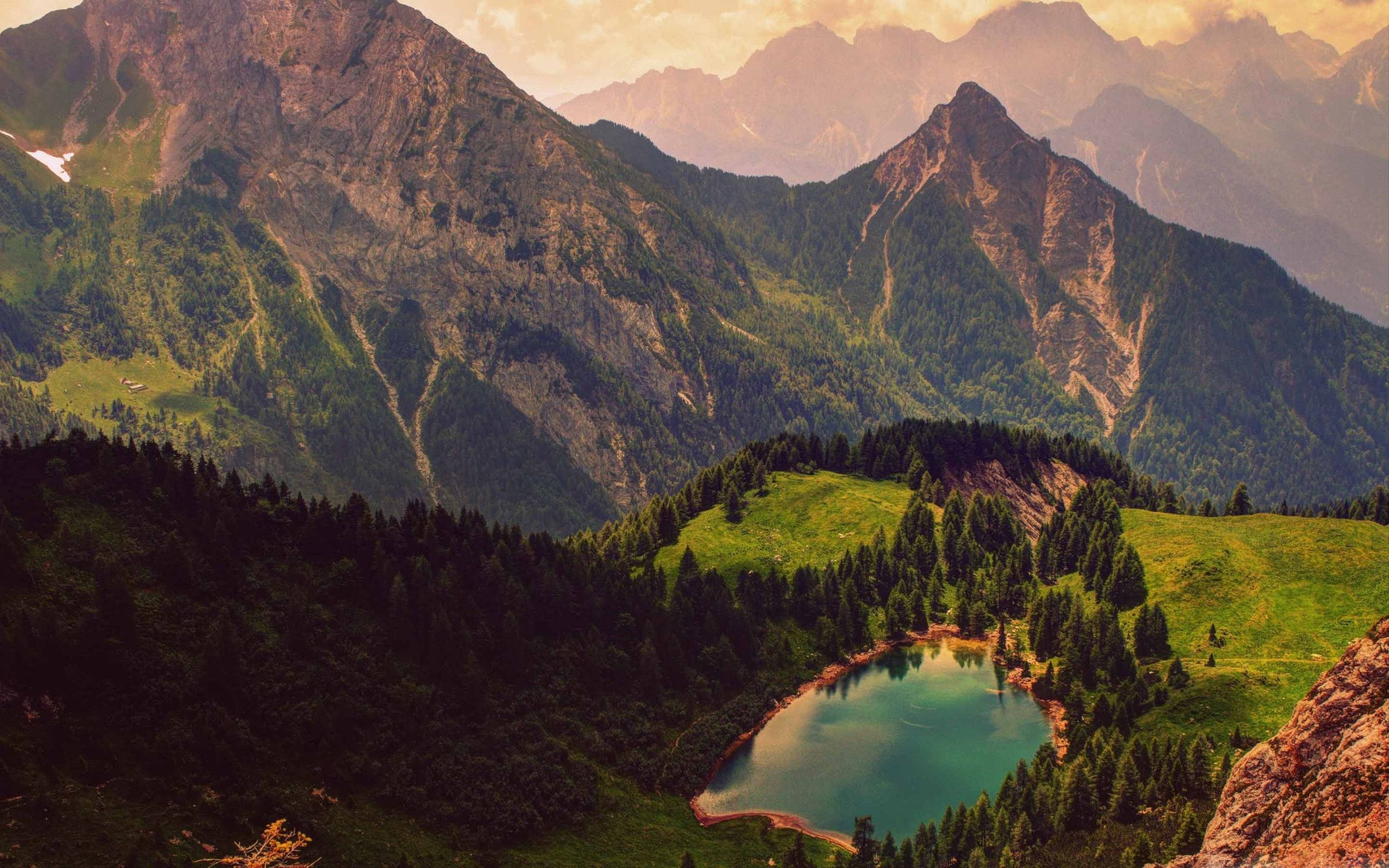 Slovenia 桌布 壁紙 — 一個寧靜的高山湖泊，坐落在鬱鬱蔥蔥的綠色山谷中，周圍環繞著高聳崎嶇的山脈，沐浴在金色的日落光芒中。