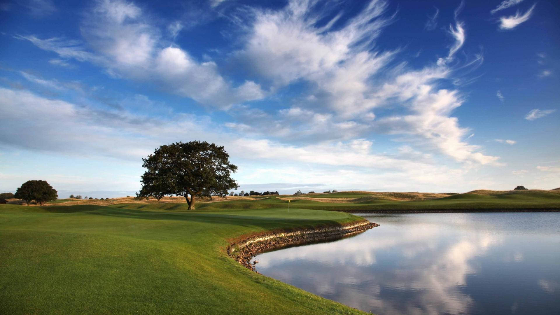 Golfing 桌布 壁紙 — 一個寧靜的高爾夫球場，擁有茂密的綠色球道、平靜的水障礙，以及在充滿活力的藍天和縷縷白雲下的一棵孤樹。