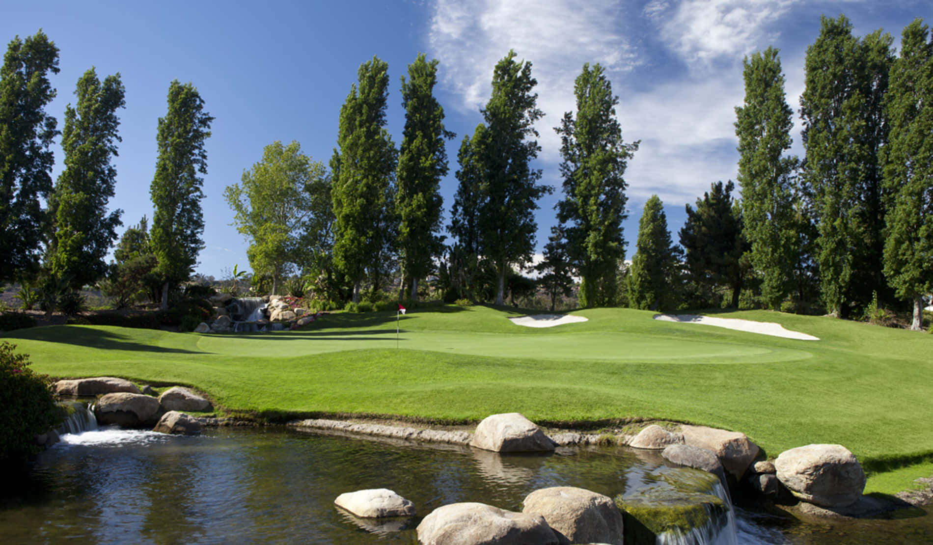 Golf Green 桌布 壁紙 — 一個寧靜的高爾夫球場果嶺，周圍環繞著高大的樹木和岩石水景，在明亮的藍天下。