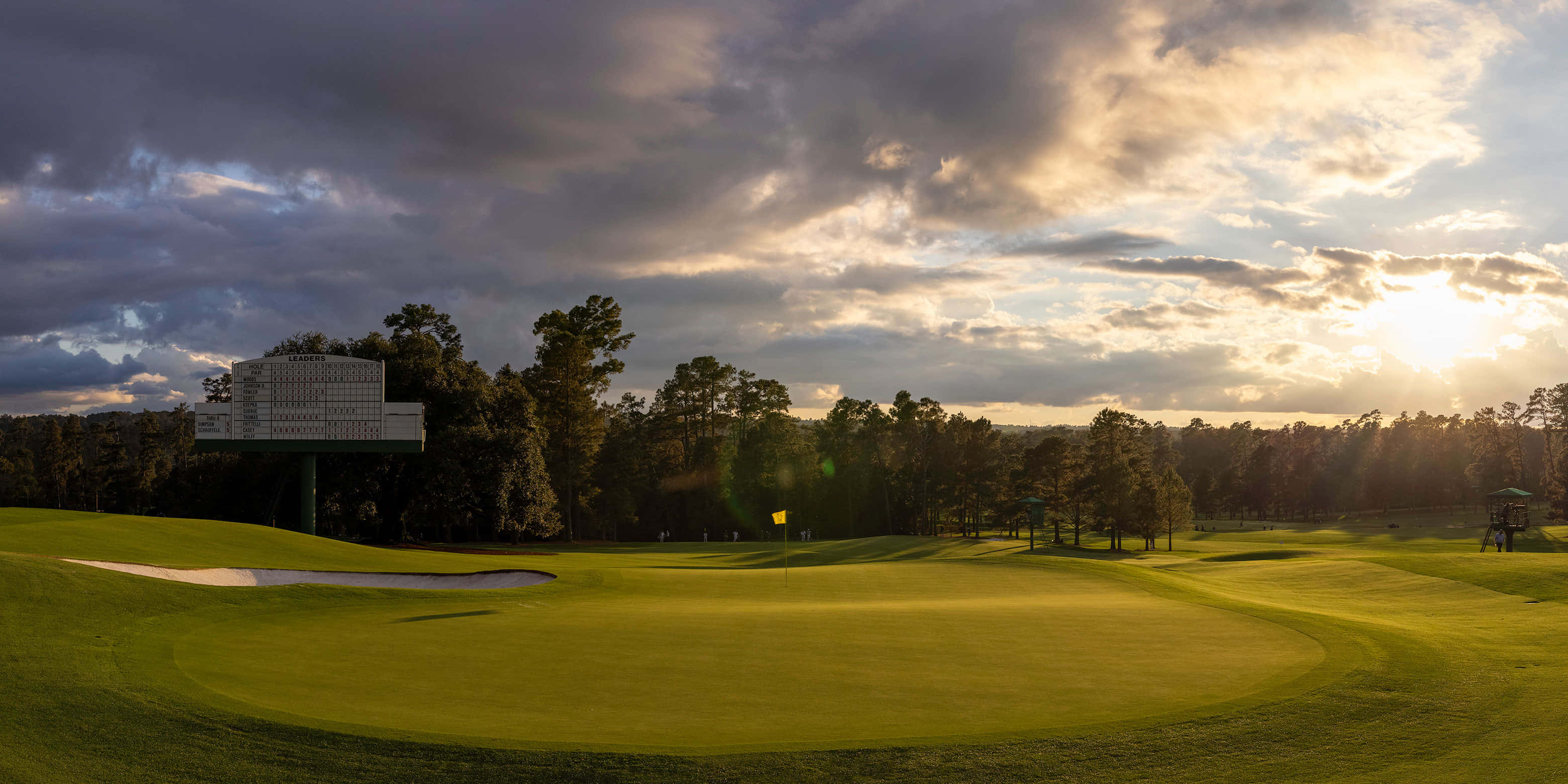 Augusta National iPhone 桌布 壁紙 — 一個寧靜的高爾夫球場沐浴在金色夕陽下，帶有戲劇性的雲彩和遠處的記分牌。