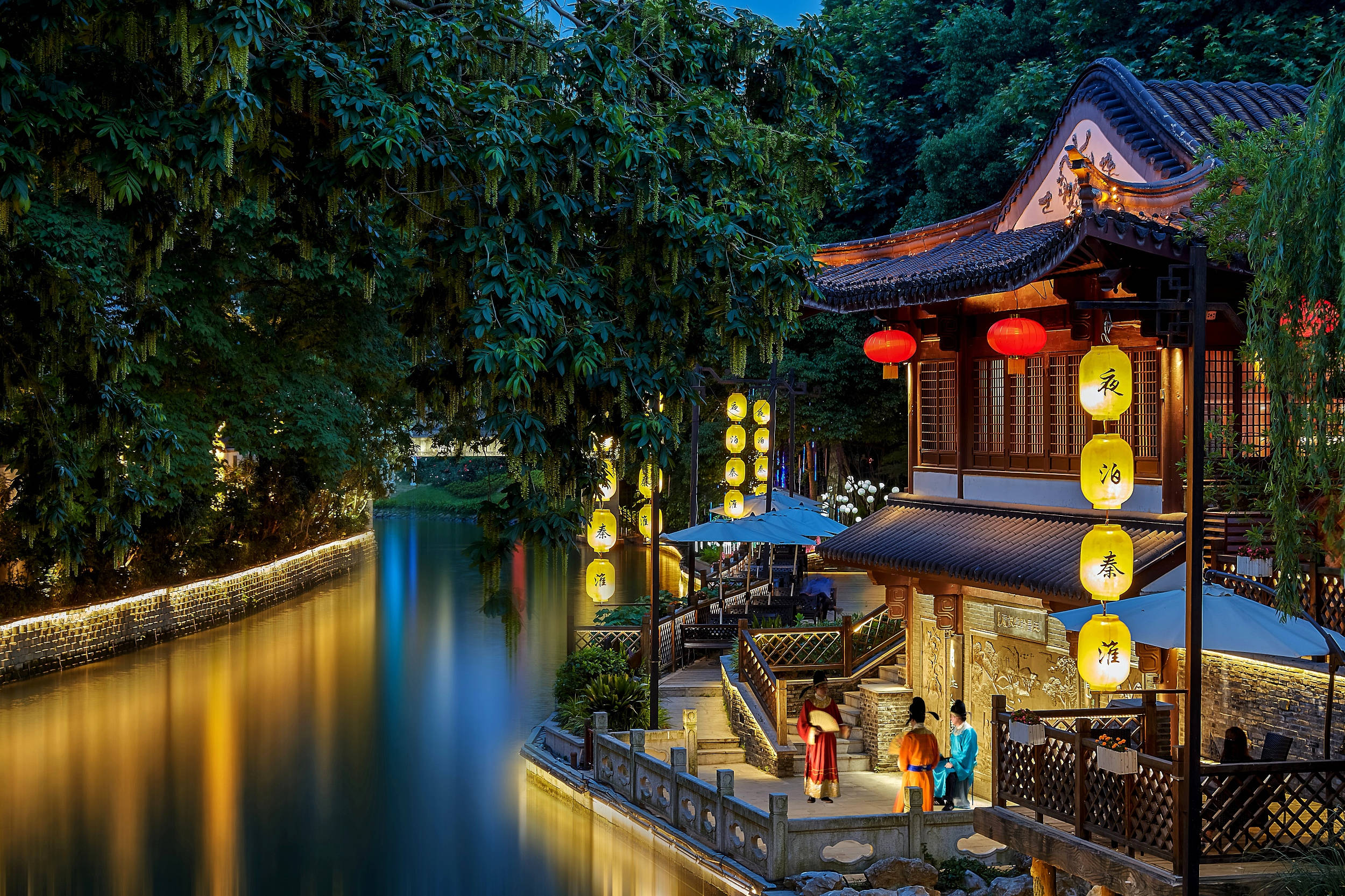 Nanjing 桌布 壁紙 — 一個寧靜的黃昏場景，傳統中式亭閣坐落在平靜的河邊，被發光的燈籠和柔和的環境光照亮。