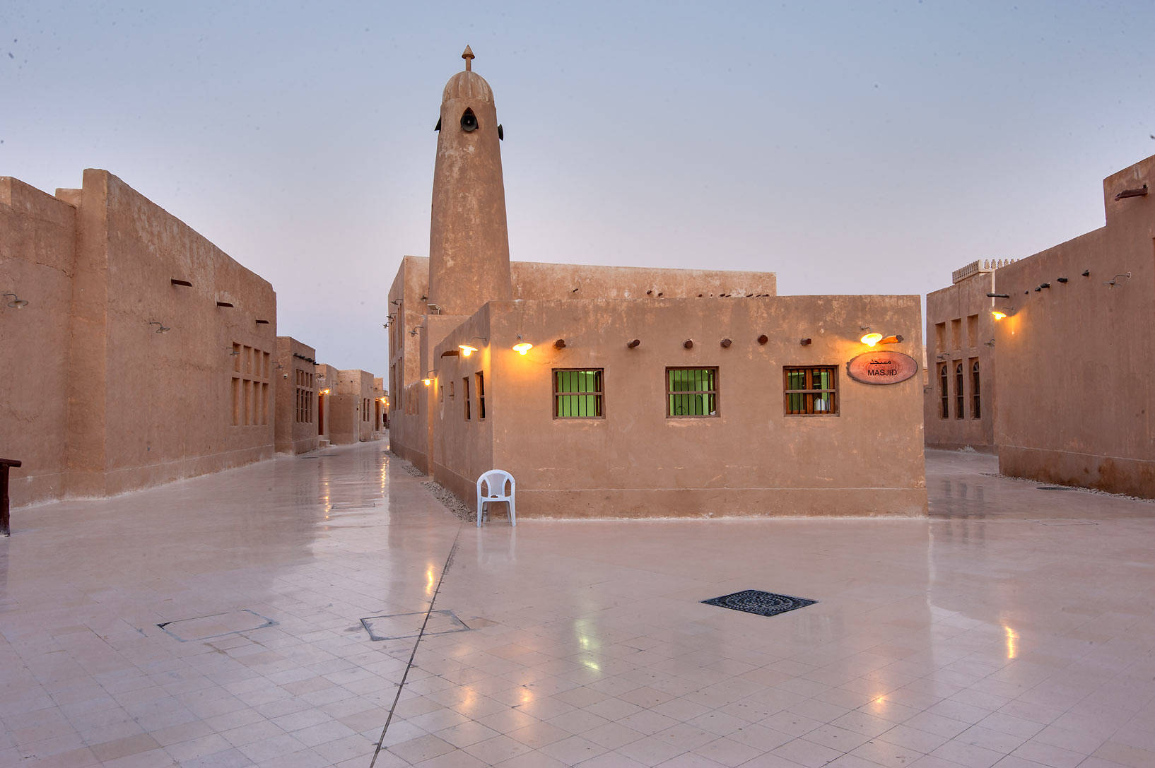 Qatar 桌布 壁紙 — 一個寧靜的黃昏庭院，有溫暖燈光的泥磚建築，以及一張孤獨的白色塑膠椅在濕石磚上反射。