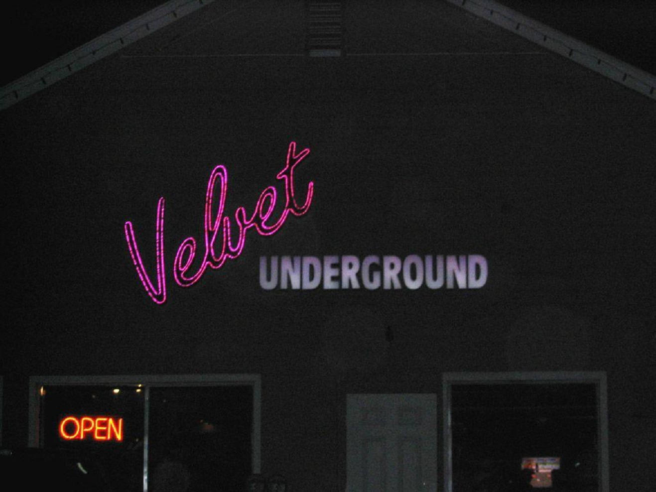 band 桌布 壁紙 — 一個寫著 Velvet Underground 的霓虹燈牌在夜間對著黑暗的建築物外牆發出粉紅色和白色的光芒，窗戶裡亮著營業中的標誌。