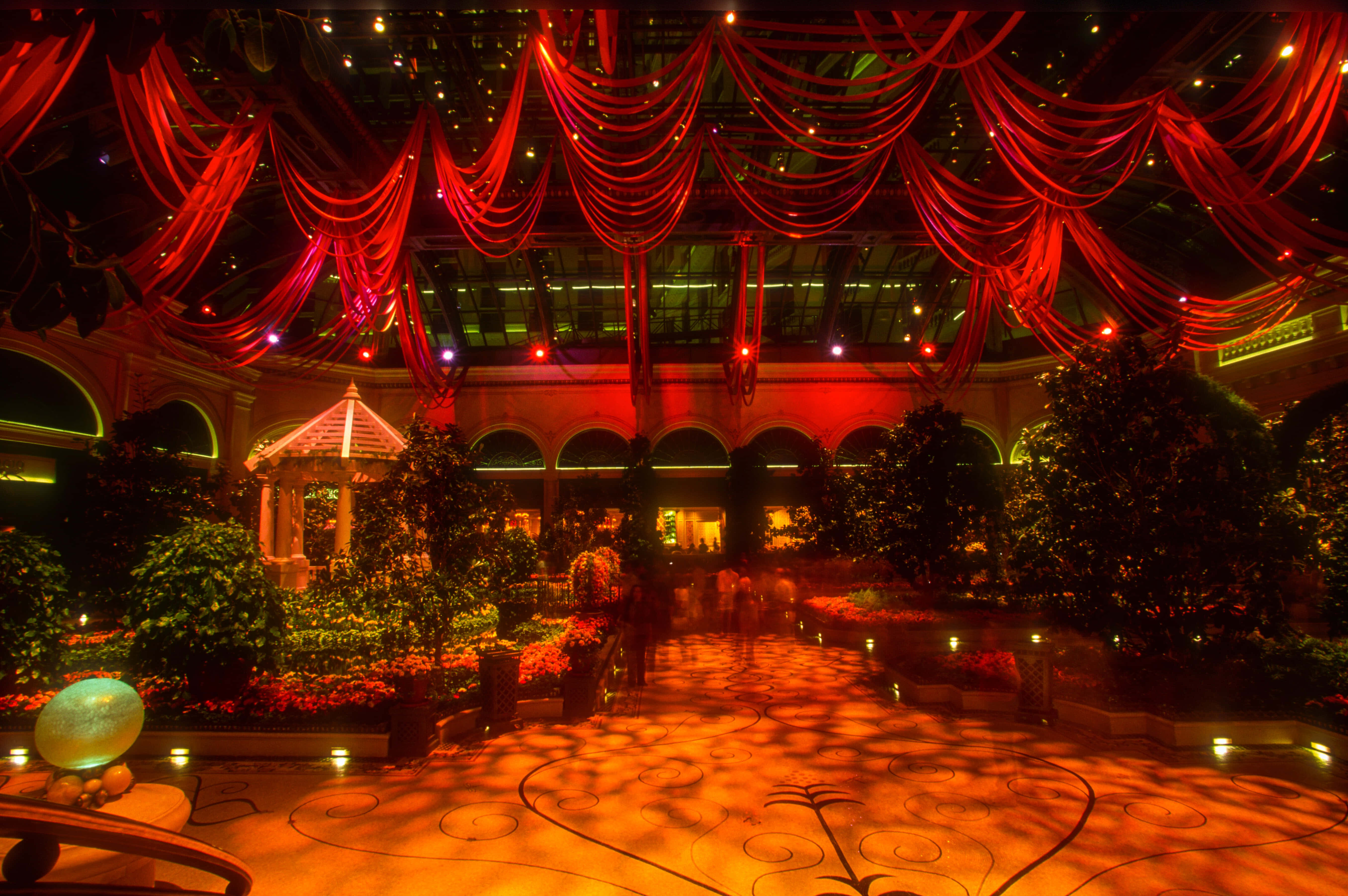 Ballroom 桌布 壁紙 — 一個寬敞的室內中庭，沐浴在溫暖的紅光中，上方有飄逸的窗簾，地板上有華麗的圖案。