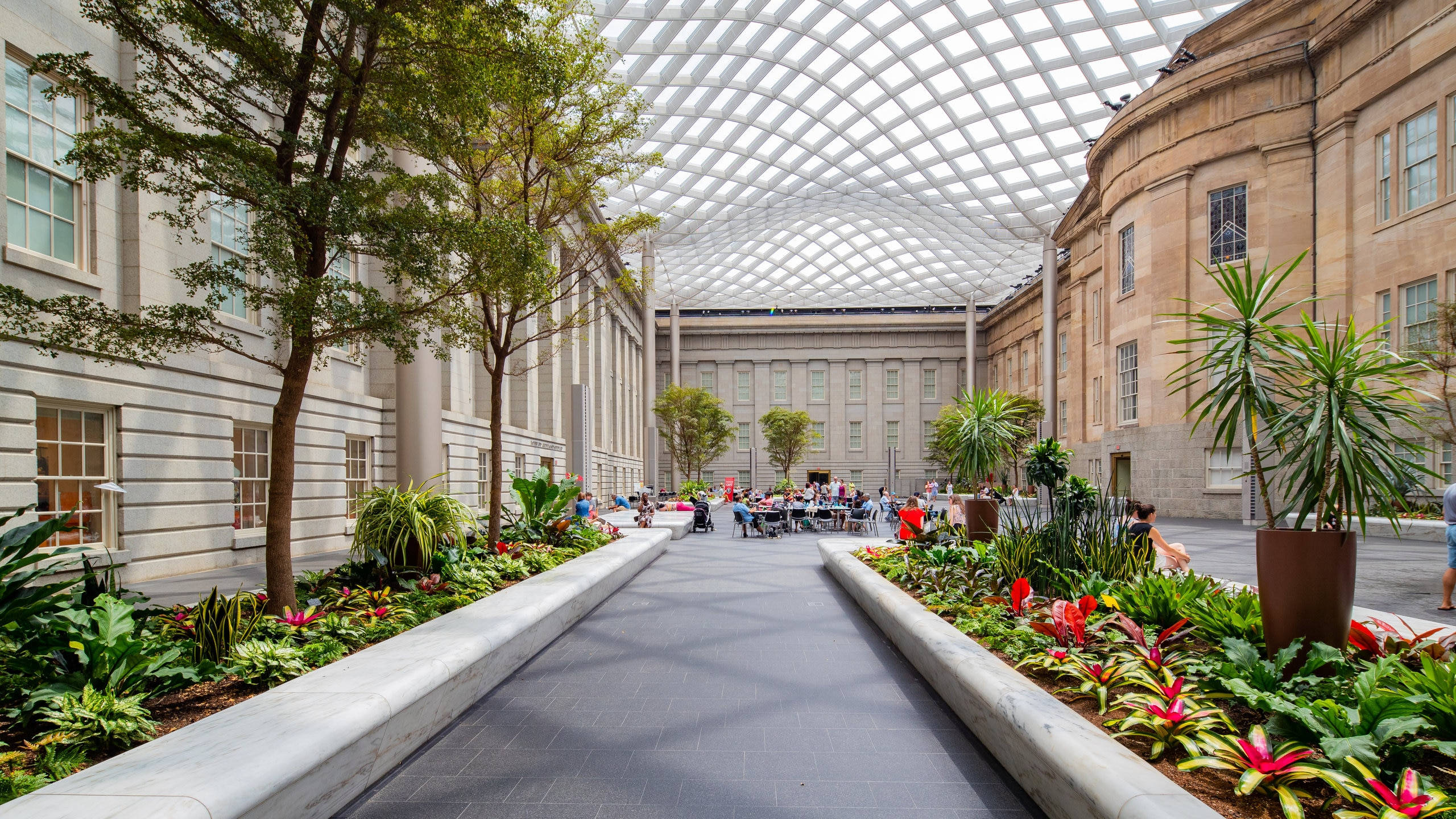 Smithsonian 桌布 壁紙 — 一個寬敞的室內庭院，擁有茂盛的熱帶植物，人們在玻璃圓頂天花板下放鬆身心。