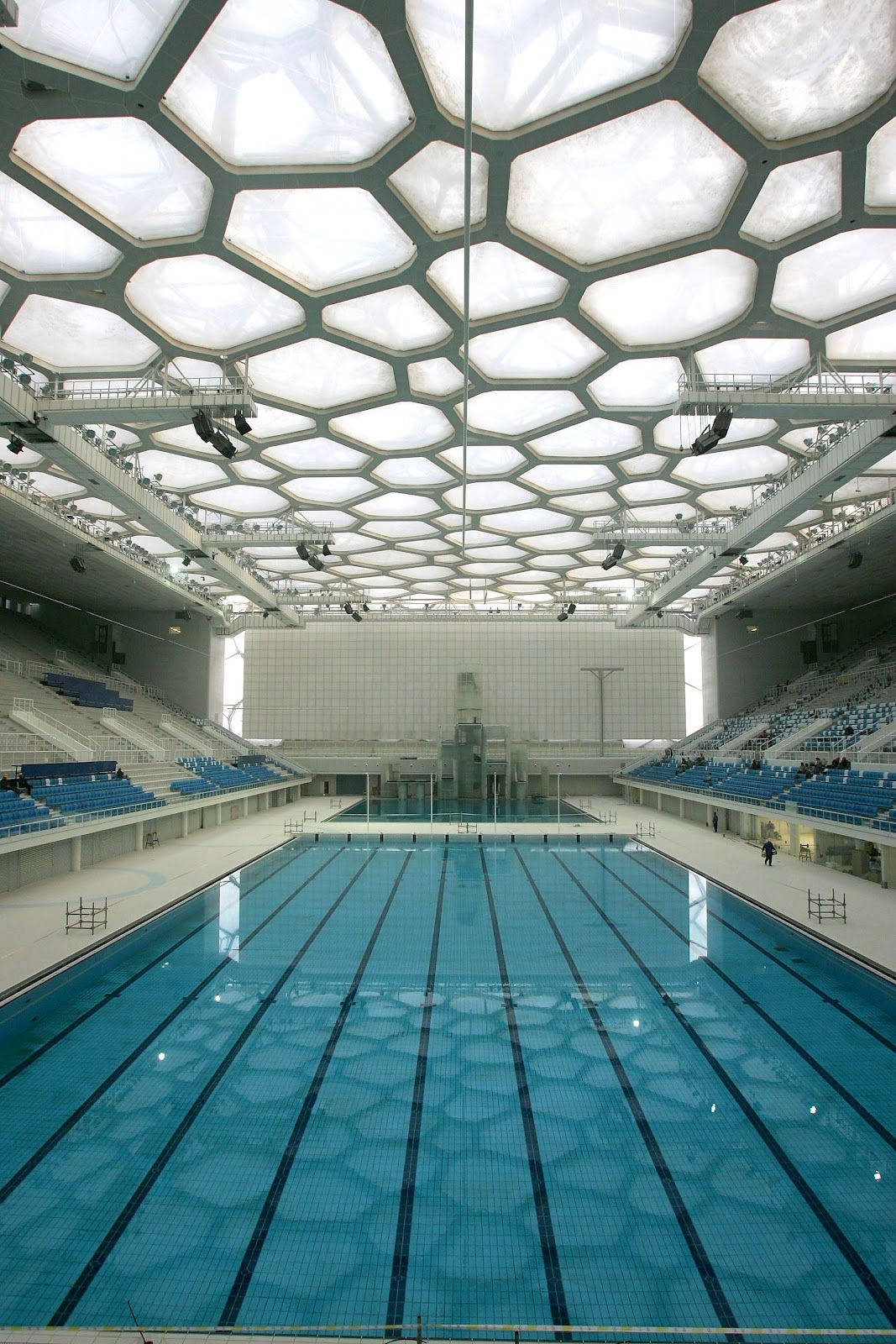 Swimming 桌布 壁紙 — 一個寬敞的室內游泳池，上方有引人注目的六角形玻璃天花板，泳道周圍設有分層藍色座位。