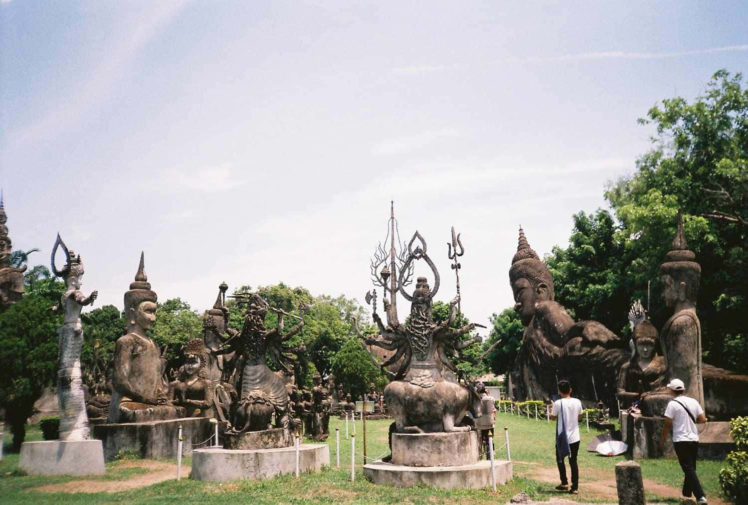 Laos 桌布 壁紙 — 一個寬敞的戶外雕塑公園，在明亮的天空下充滿了大型風化的神靈和神話人物雕像。