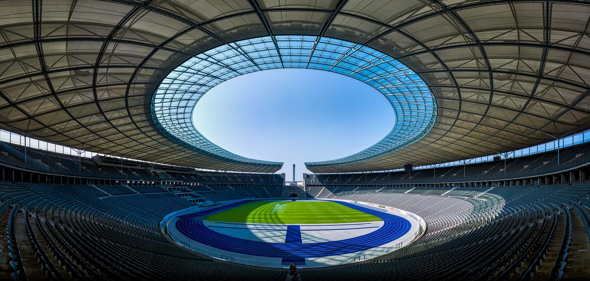 Stadium 桌布 壁紙 — 一個寬敞的現代化體育場，設有圓形玻璃屋頂和空藍色座位，圍繞著綠色跑道和場地。