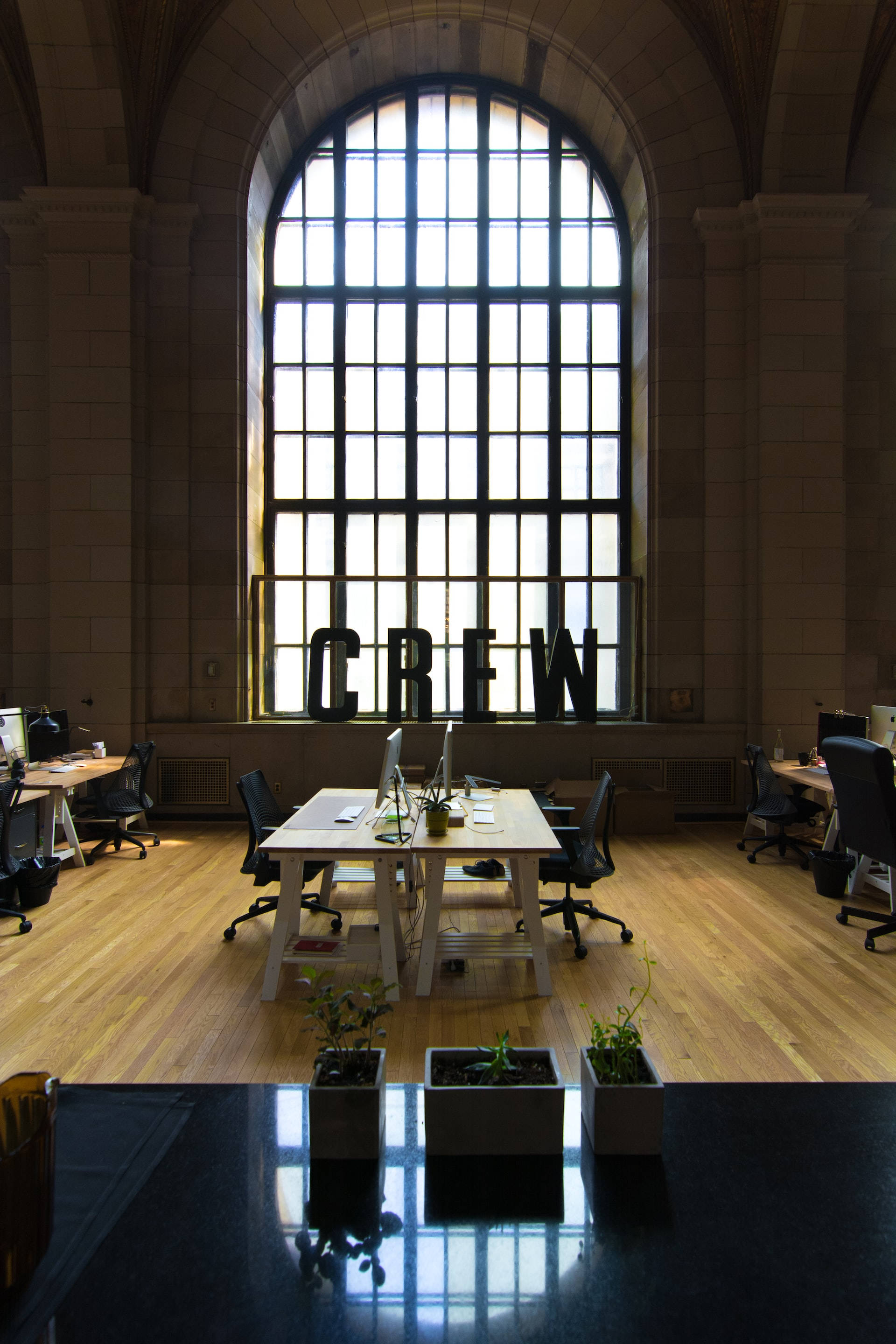 Office Space 桌布 壁紙 — 一個寬敞的石牆辦公室，鋪有木地板，巨大的拱形窗戶將光線投射在辦公桌和「CREW」標誌上。