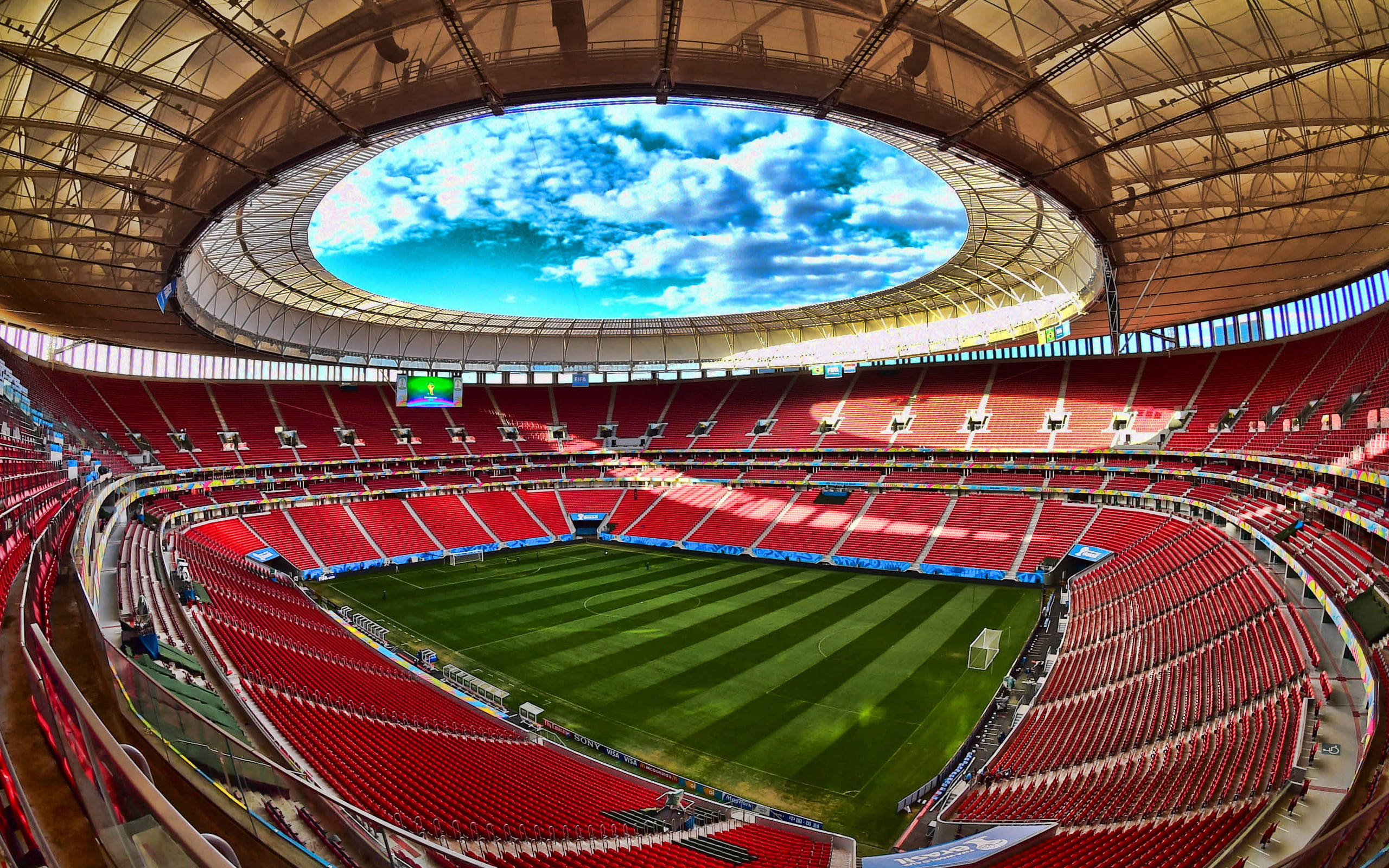 Stadium 桌布 壁紙 — 一個寬敞的空足球場，設有紅色座位和綠色草坪，圓形屋頂下露出明亮的藍天和蓬鬆的白雲。