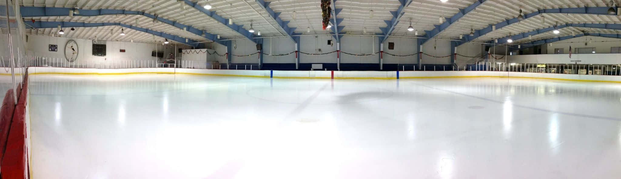 Ice Rink 桌布 壁紙 — 一個寬敞空曠的室內溜冰場，明亮的白色冰面反射著頭頂燈光和藍色邊框的牆壁。