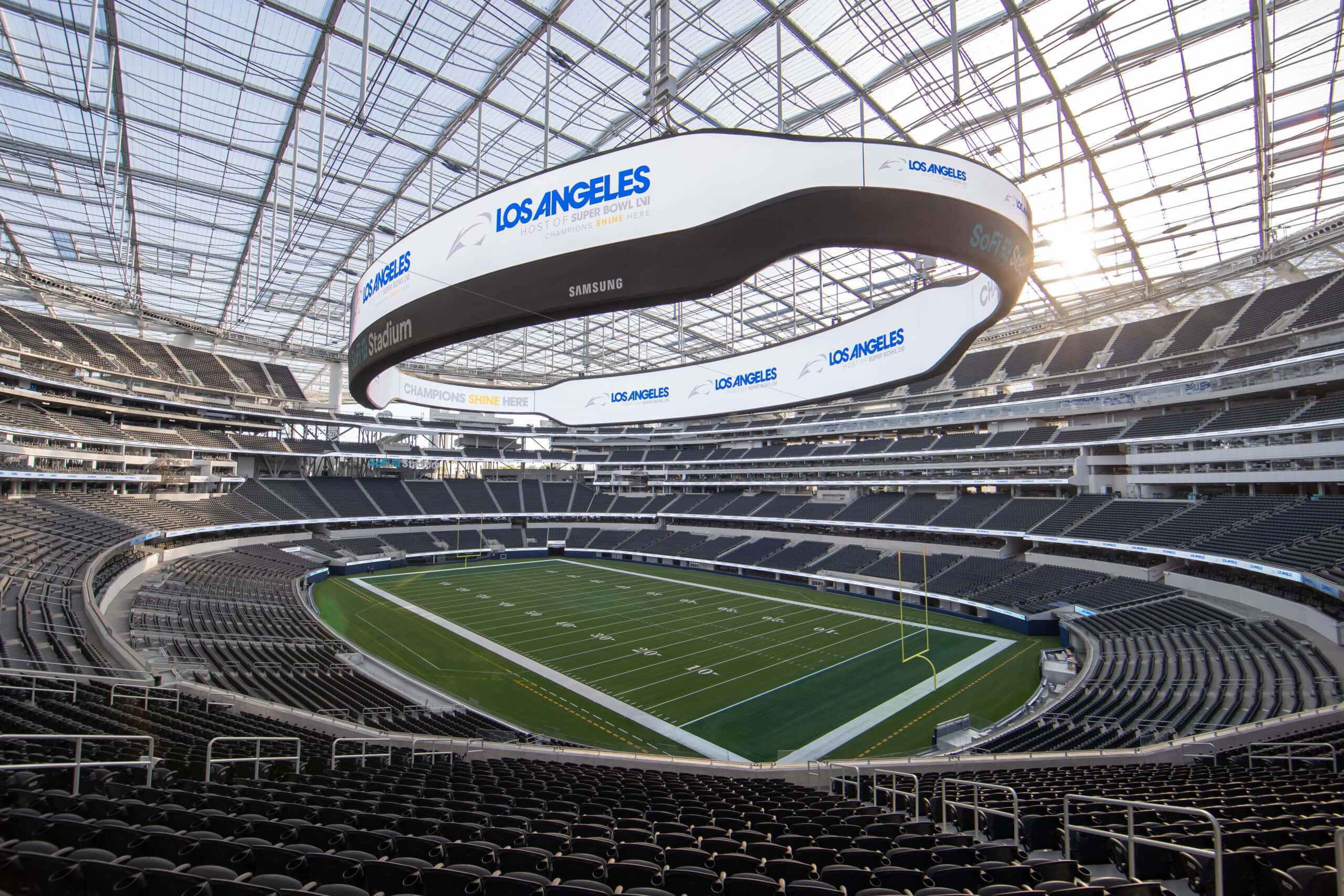 Sofi Stadium 桌布 壁紙 — 一個寬敞空曠的足球場，設有玻璃屋頂和「洛杉磯」標誌，沐浴在明亮的日光下。
