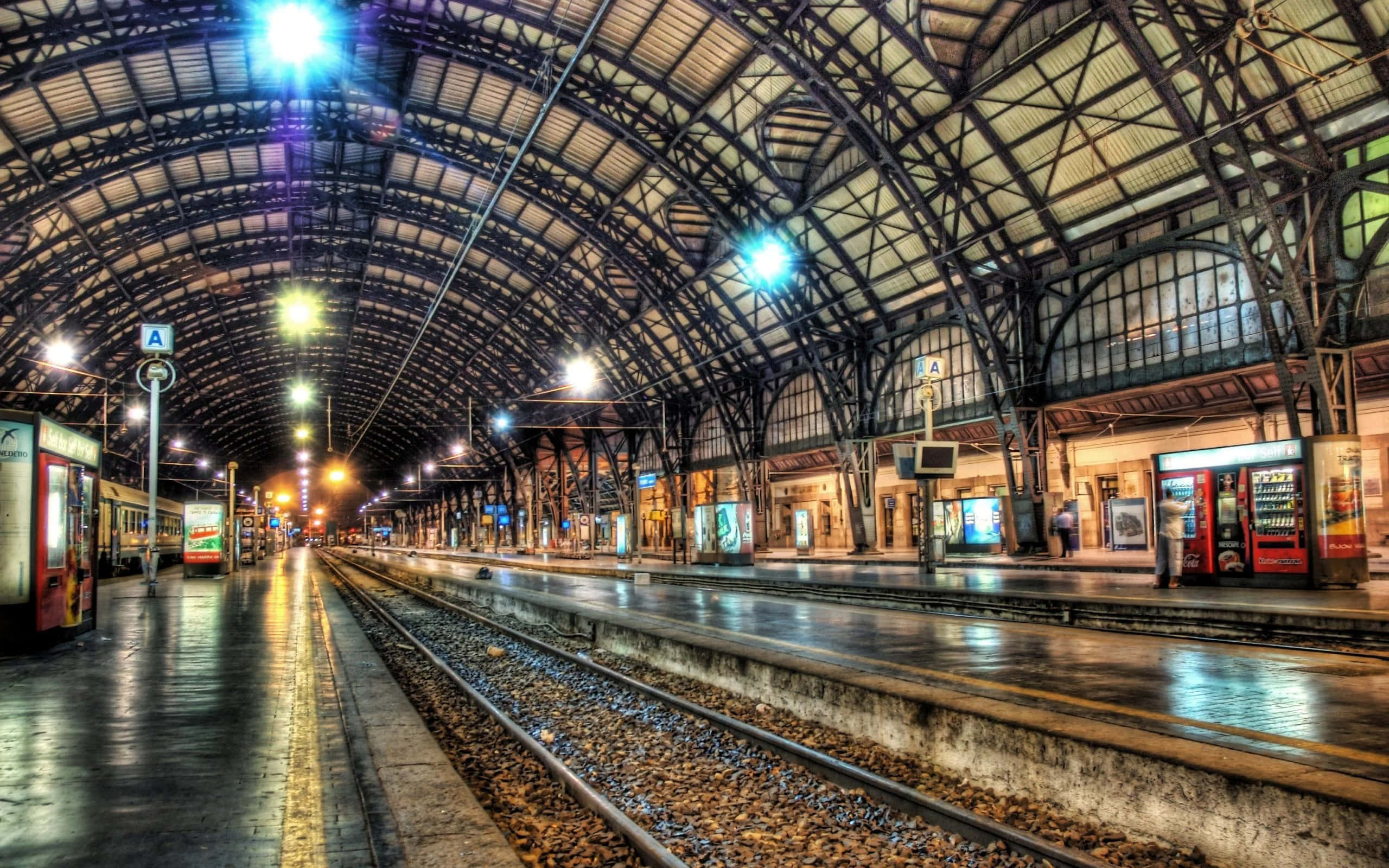 Train Station 桌布 壁紙 — 一個寬闊的拱形火車站，潮濕的月台在巨大金屬屋頂下反射出鮮豔燈光。