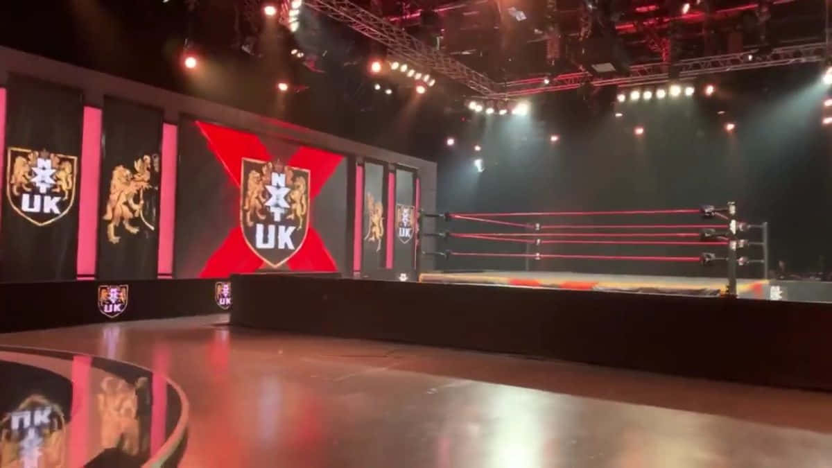 Wrestling Ring 桌布 壁紙 — 一個專業摔角擂台，在戲劇性的紅色舞台燈光下，周圍螢幕顯示NXT UK品牌。