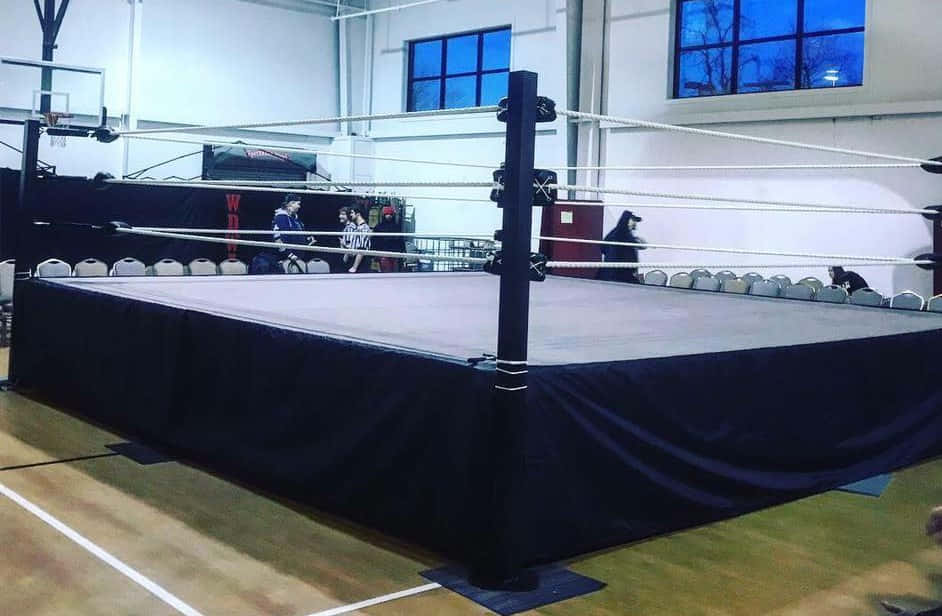 Wrestling Ring 桌布 壁紙 — 一個專業摔角擂台，黑色帆布和繩索，位於體育館中央，在人工燈光下。