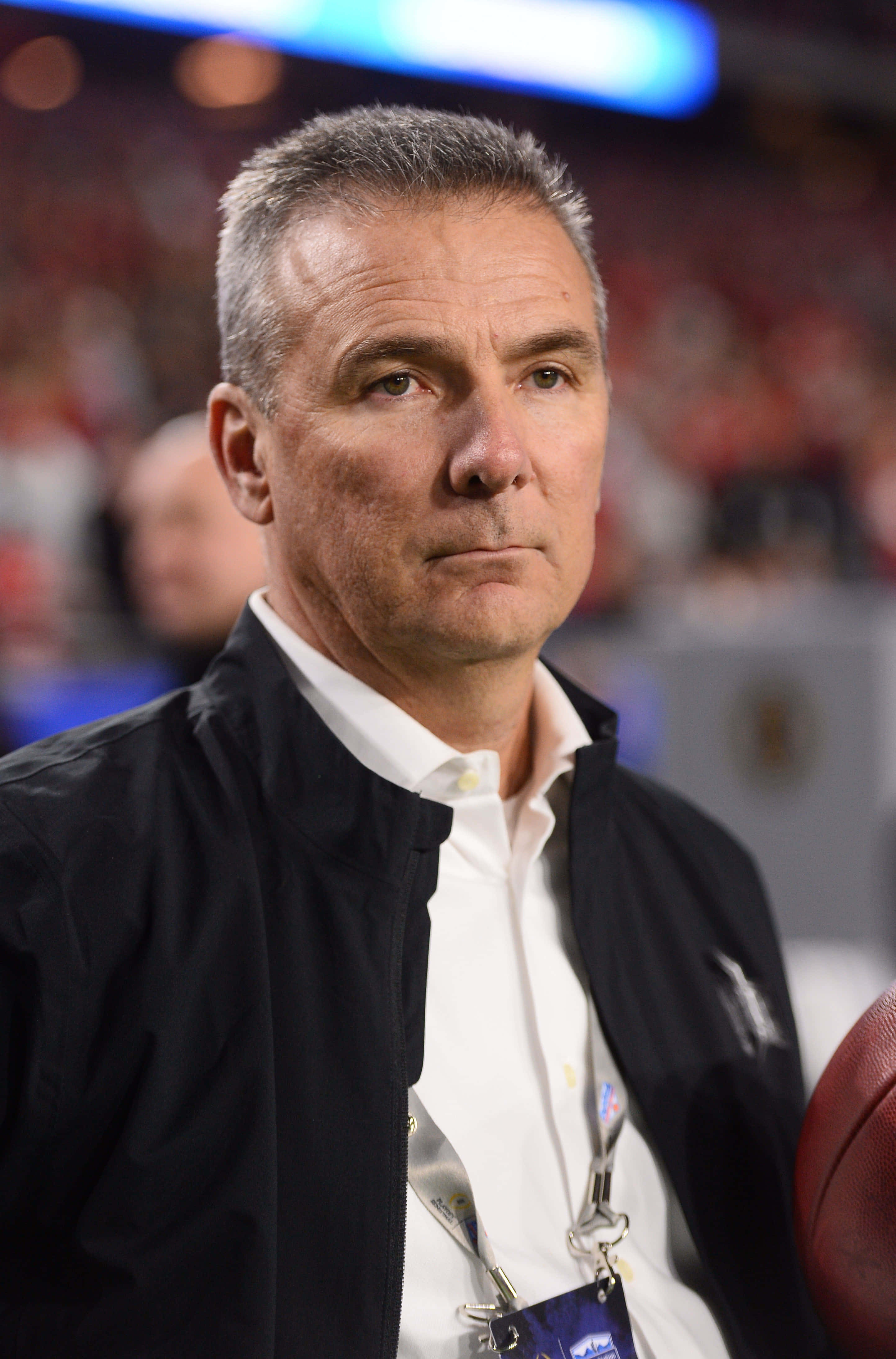Urban Meyer 桌布 壁紙 — 一個專注的中年男子穿著黑色外套，在燈光明亮的體育館裡拿著一個足球，身後是模糊的紅色座位。