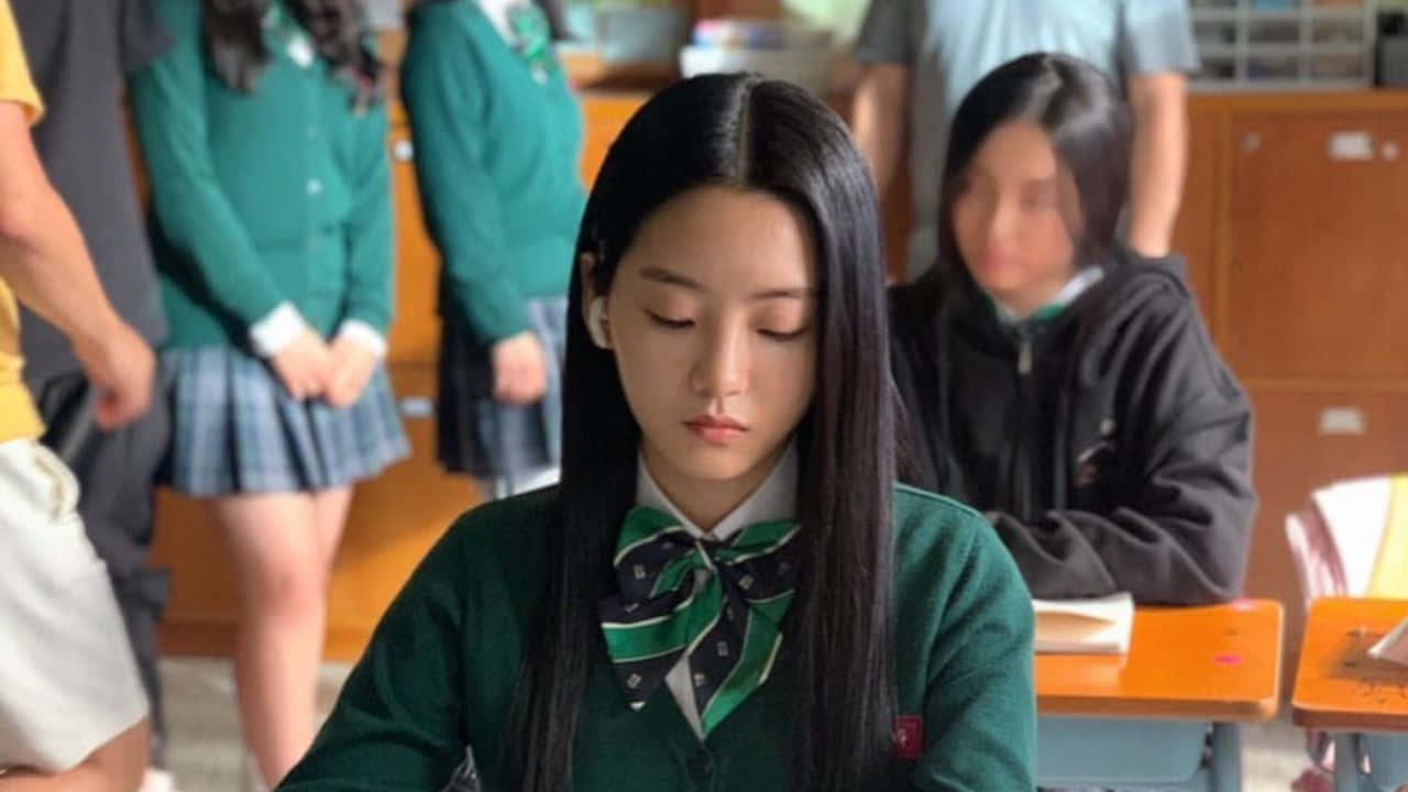 All Of US Are Dead 桌布 壁紙 — 一個專注的十幾歲女孩穿著綠色校服，坐在教室的書桌前，背景有同學。