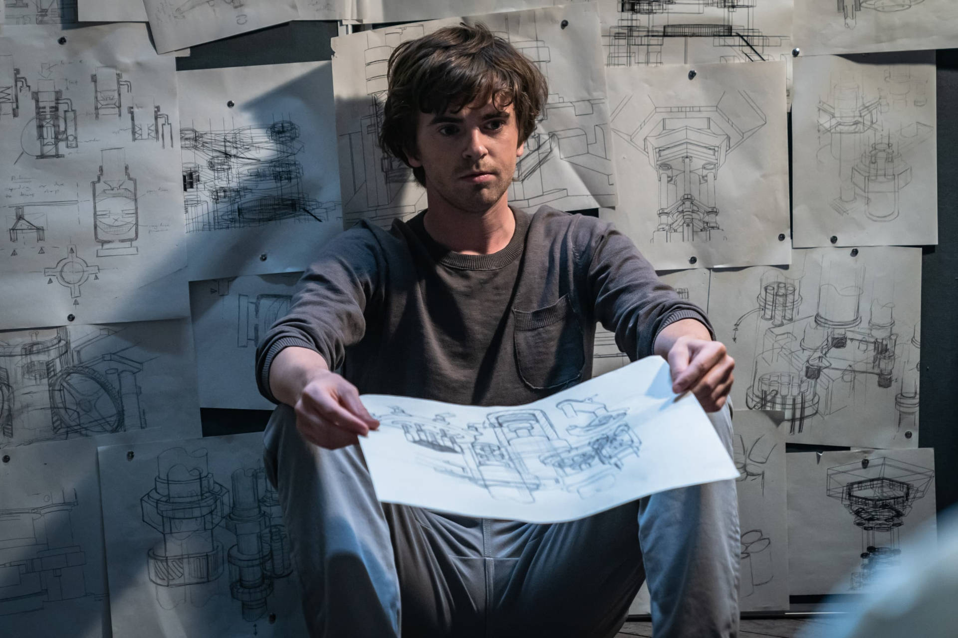 Freddie Highmore 桌布 壁紙 — 一個專注的年輕人在昏暗的房間裡，舉起一張技術草圖，周圍牆上釘滿了工程圖紙。