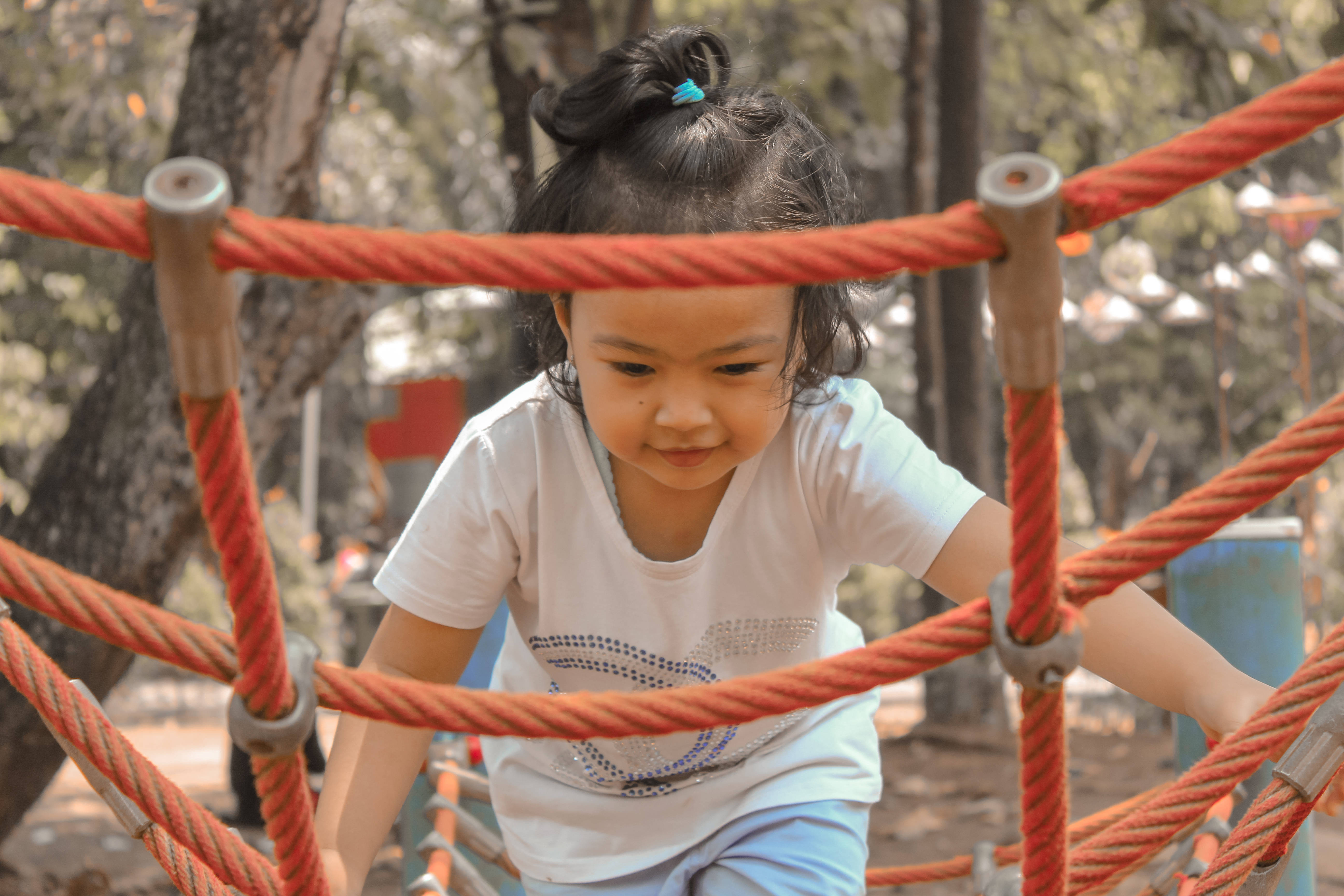 Playground 桌布 壁紙 — 一個專注的年輕女孩穿著白色襯衫，在陽光明媚的戶外遊樂場攀爬明亮的橙色繩索結構。