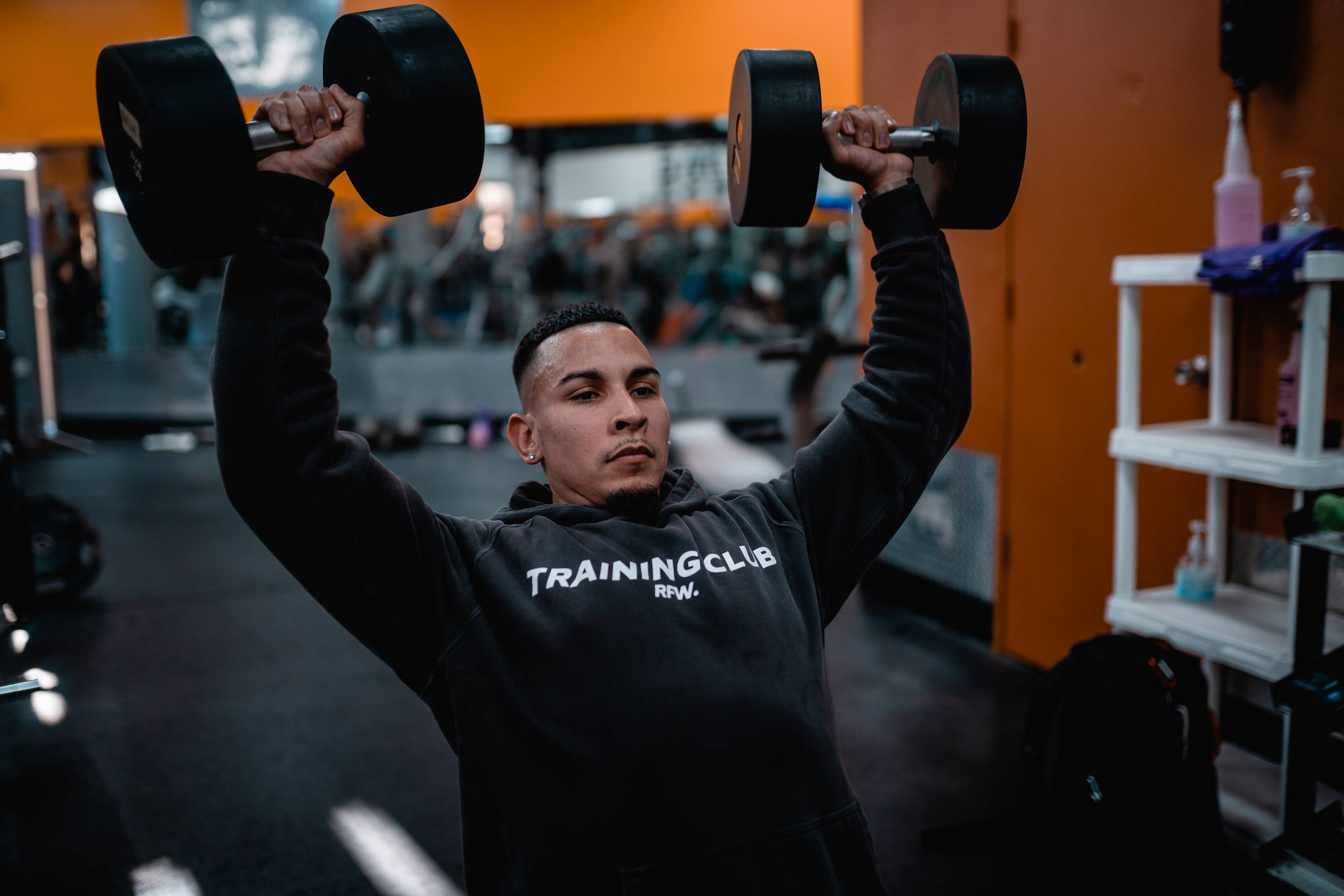 Weightlifting 桌布 壁紙 — 一個專注的年輕男子在光線充足的健身房中，將沉重的啞鈴舉過頭頂，健身房有橙色牆壁和鏡面設備。