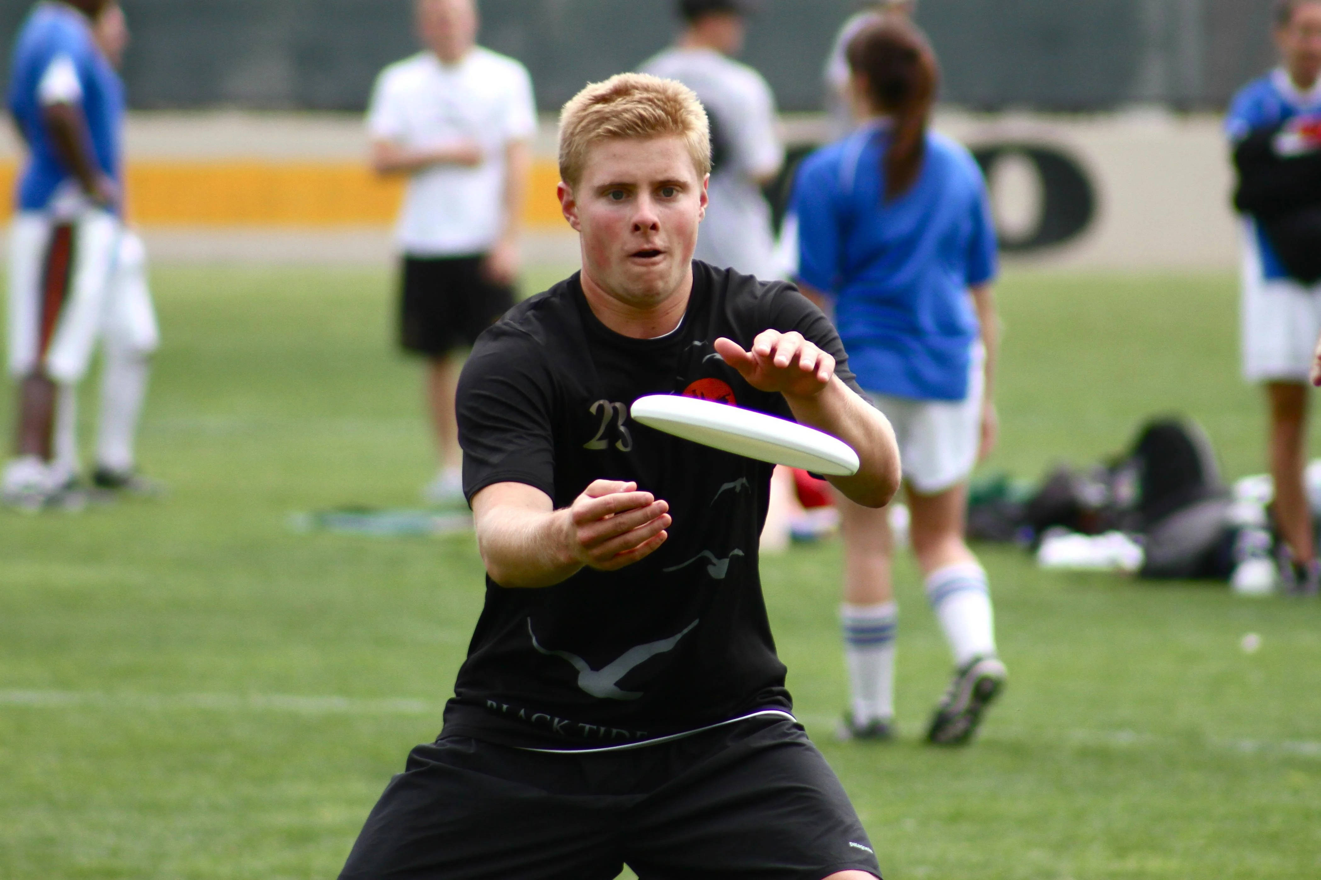 Ultimate Frisbee 桌布 壁紙 — 一個專注的年輕男子穿著黑色運動裝備，在充滿活力的綠色運動場上投擲白色飛盤，背景中的隊友模糊不清。