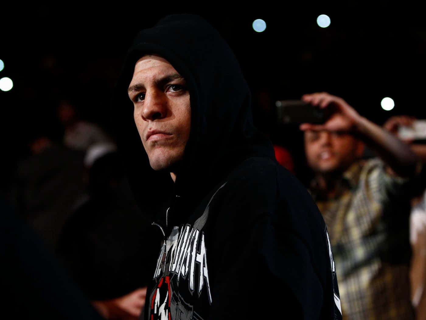 UFC 桌布 壁紙 — 一個專注的戰士穿著黑色連帽衫，在明亮的競技場燈光下凝視，身後有模糊的觀眾。
