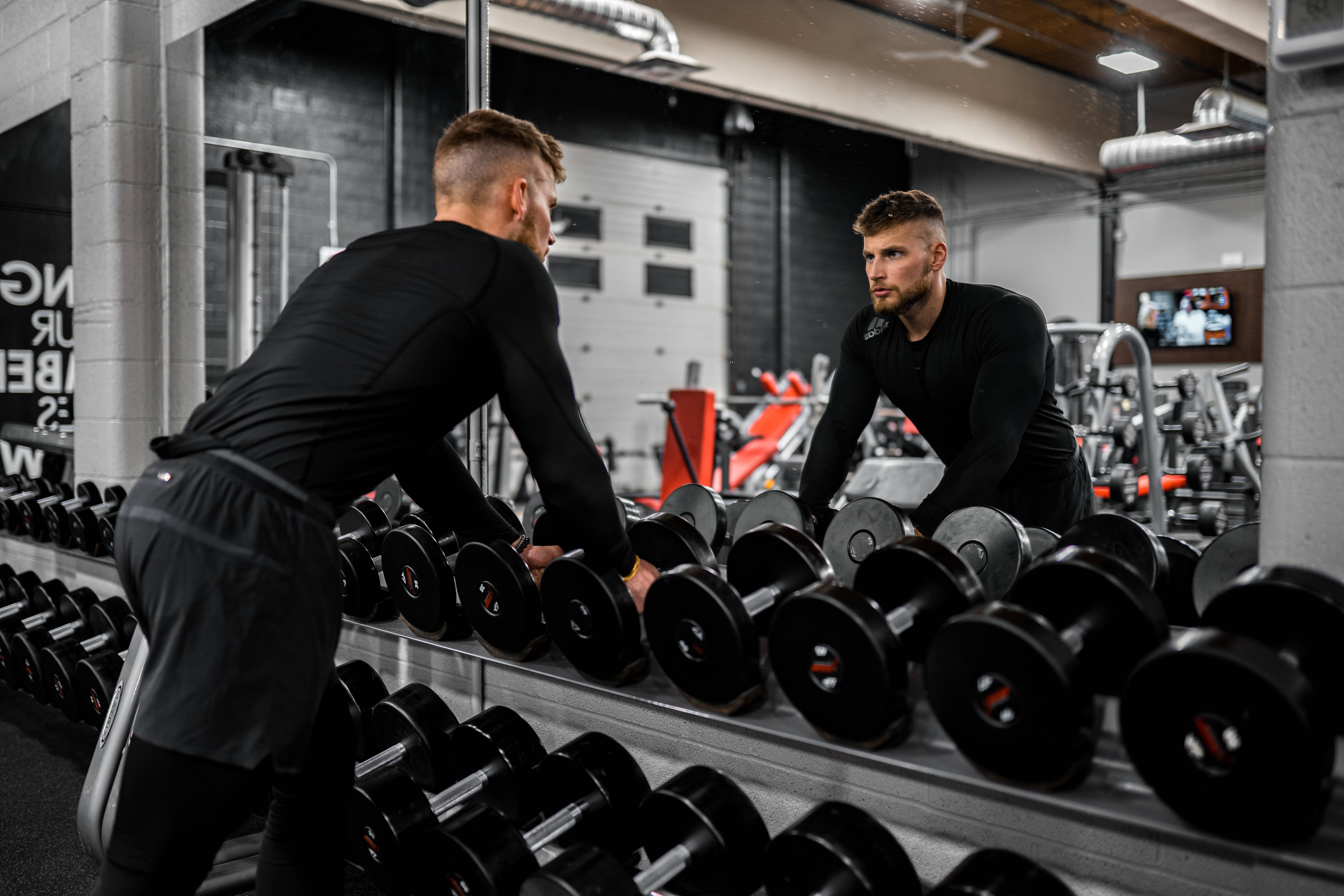 Weightlifting 桌布 壁紙 — 一個專注的男人在健身房從架上挑選啞鈴，而他的鏡中倒影正凝視回望。