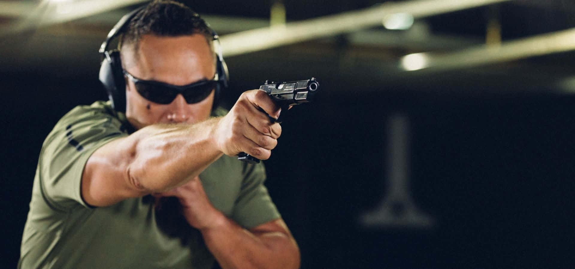 Shooting 桌布 壁紙 — 一個專注的男人戴著太陽眼鏡和耳罩，在光線昏暗的室內射擊場精確地瞄準手槍。