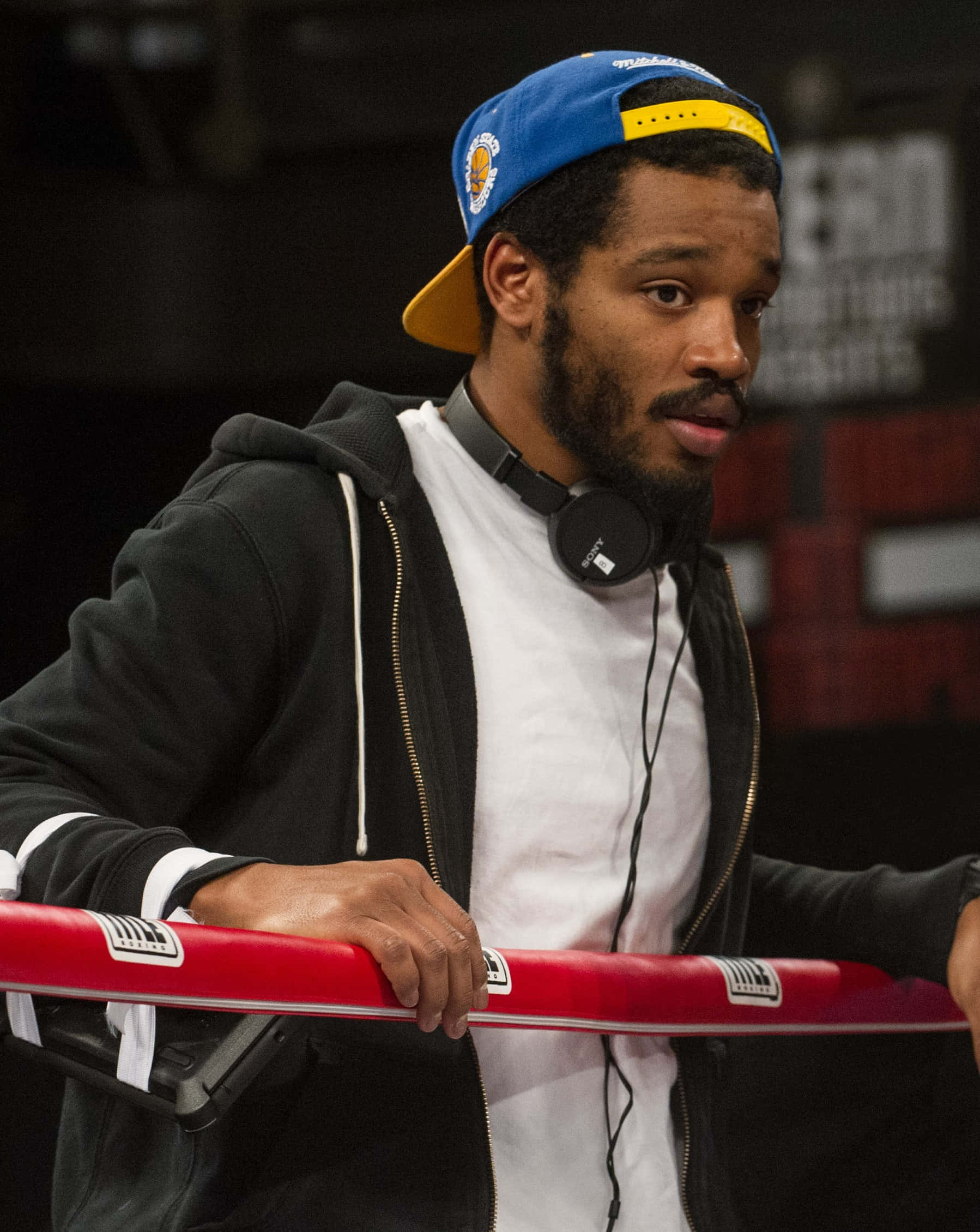 Ryan Coogler 桌布 壁紙 — 一個專注的男人戴著藍色帽子和黑色連帽衫，頸上掛著耳機，靠在紅色拳擊台繩上。