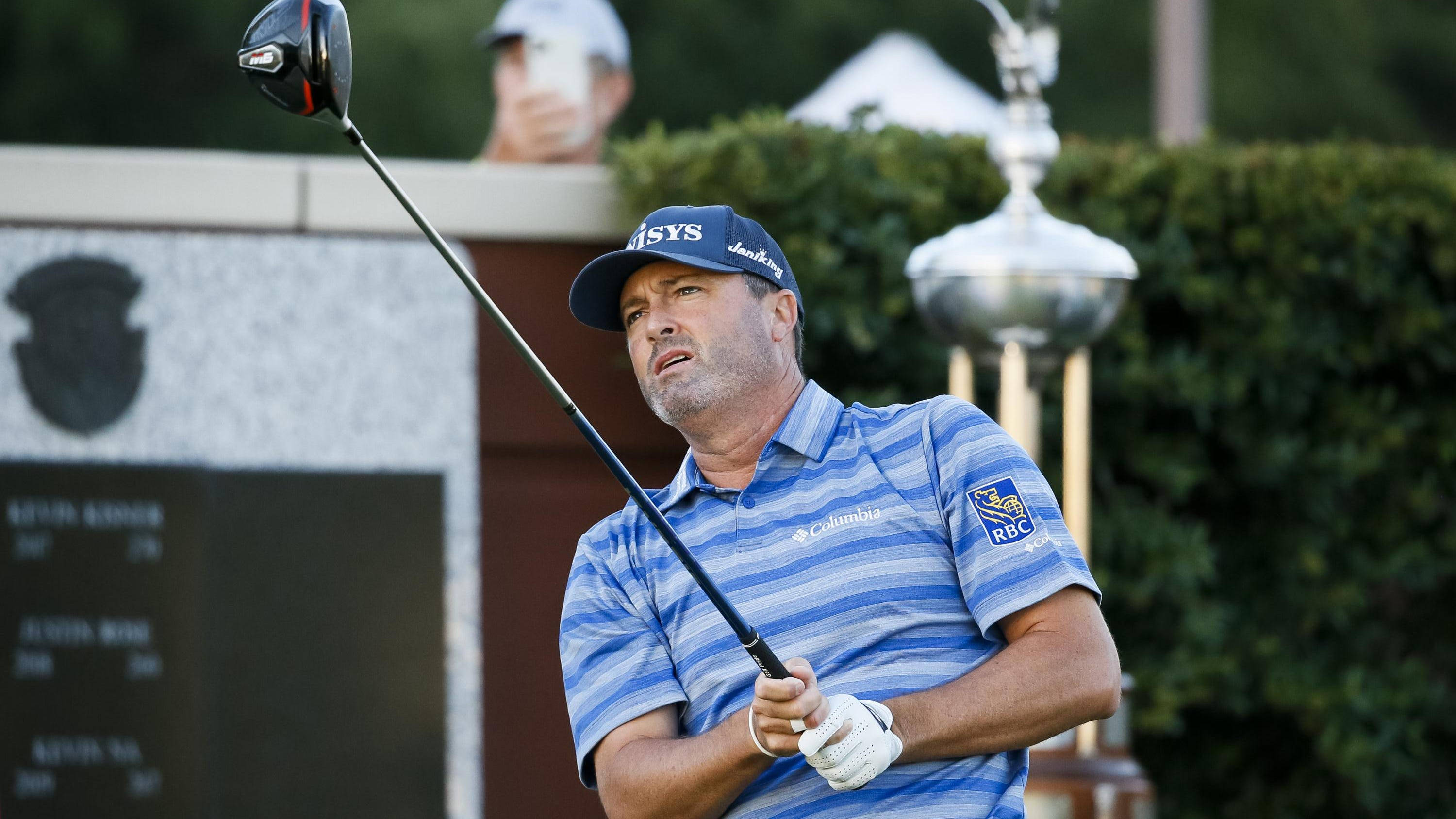 Ryan Palmer 桌布 壁紙 — 一個專注的男性高爾夫球手在揮桿中途，背景中可見一個獎盃，在陽光明媚的日子。