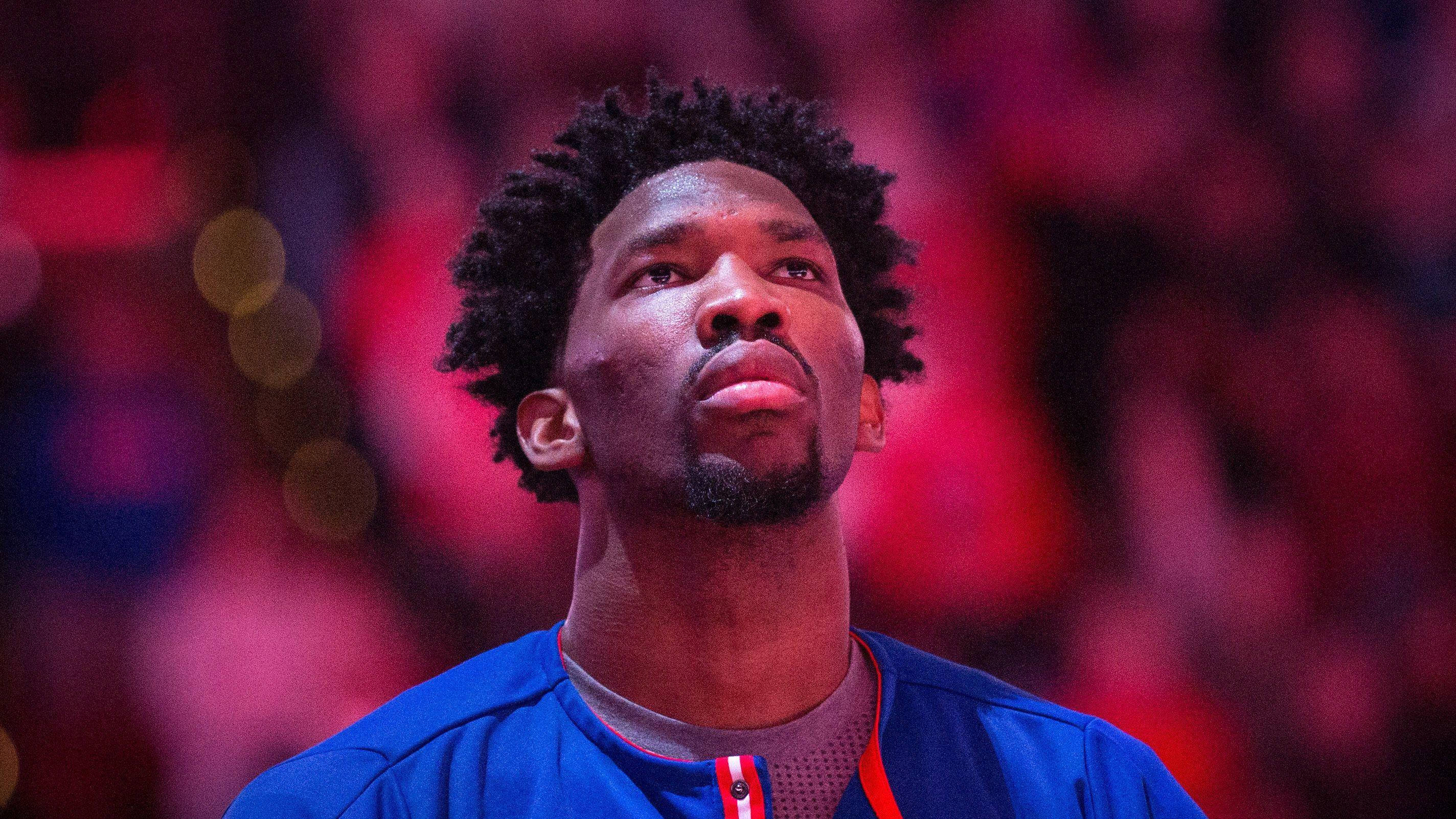 Embiid 桌布 壁紙 — 一個專注的籃球運動員，捲髮，在模糊的紅色場館背景和散景燈光中向上凝視。