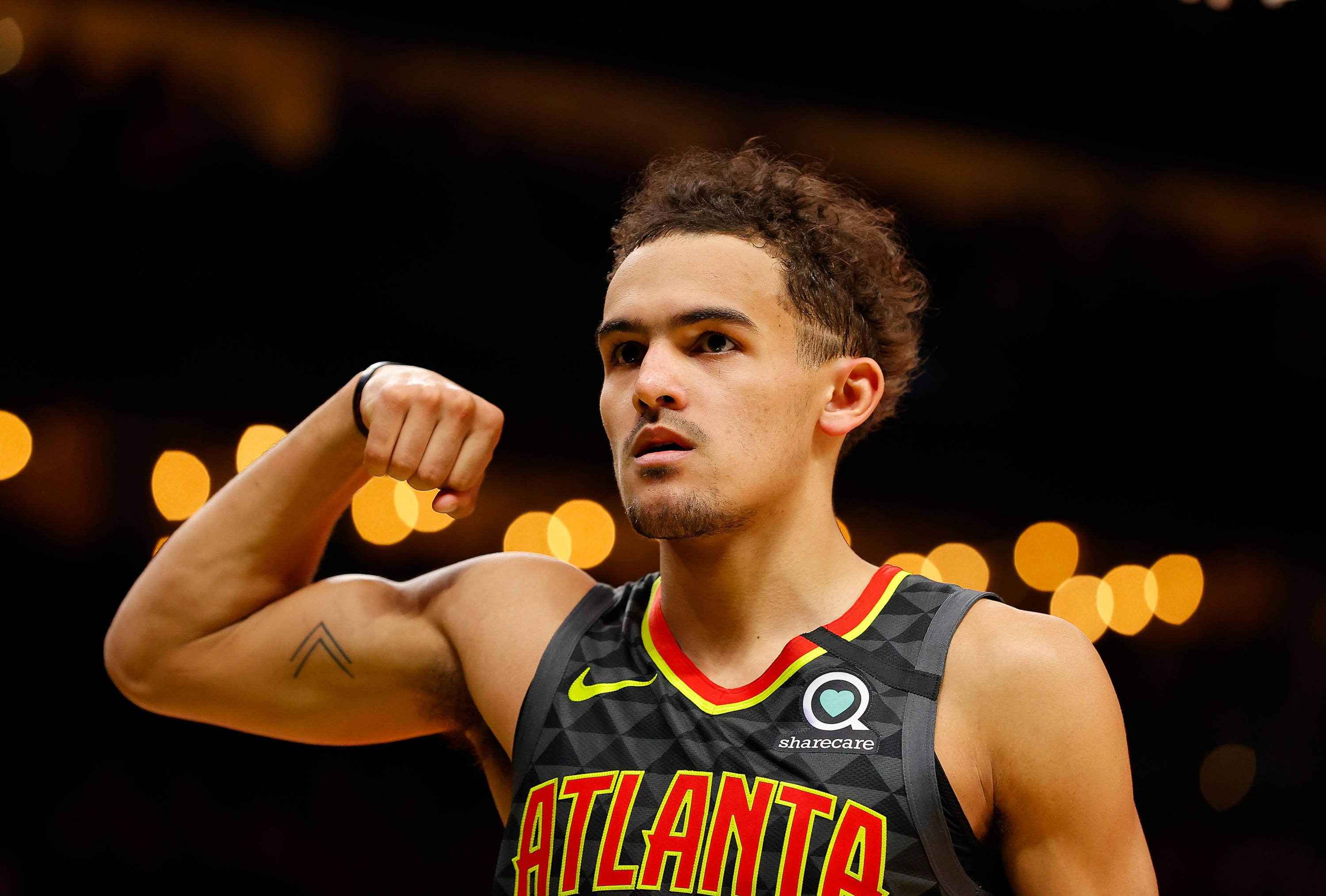Trae Young 桌布 壁紙 — 一個專注的籃球運動員在場館燈光下彎曲他的二頭肌，背景有模糊的金色散景。