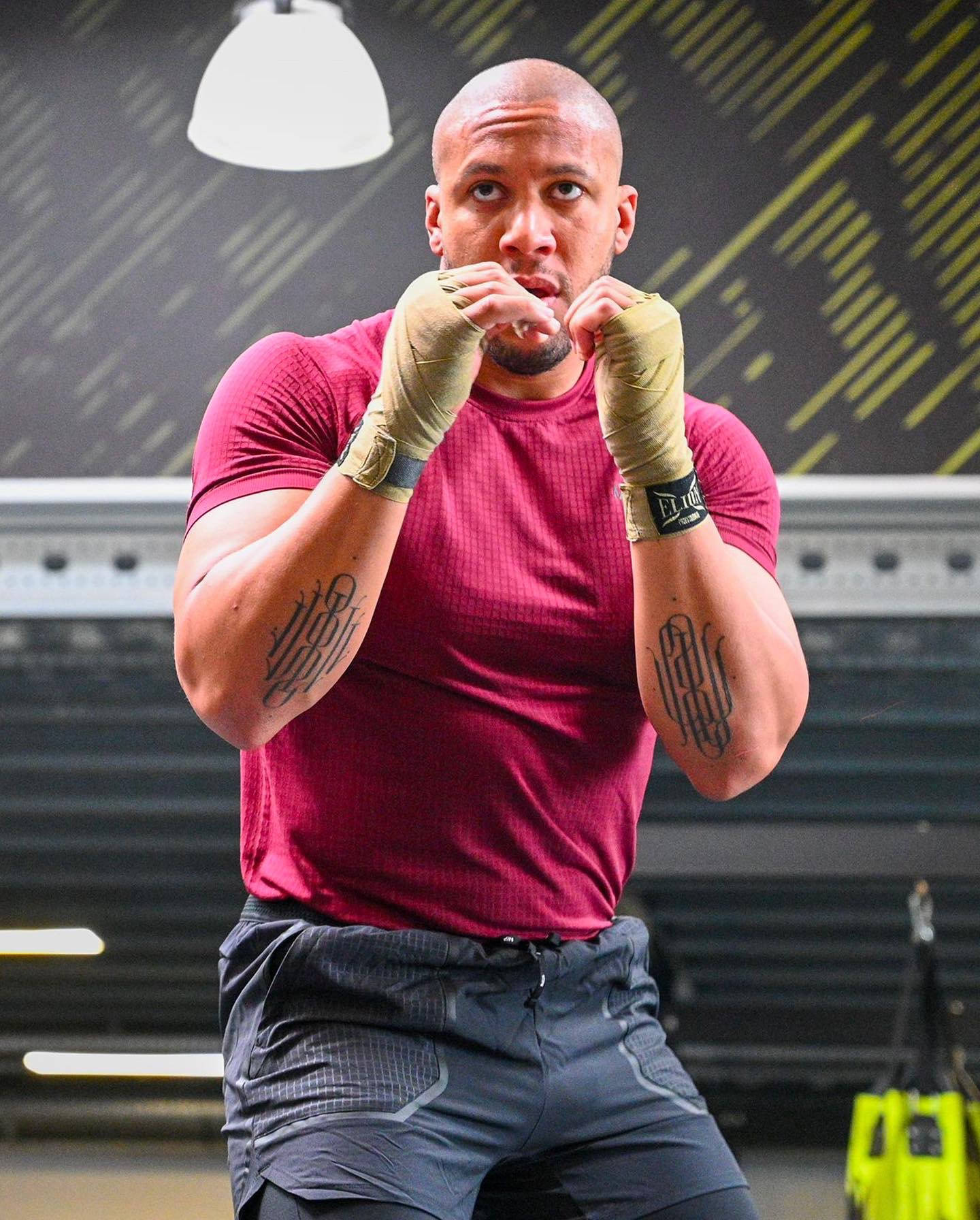 MMA 桌布 壁紙 — 一個專注的肌肉男，身穿紅色襯衫和纏手帶，在幾何牆壁圖案的健身房內擺出戰鬥姿勢。