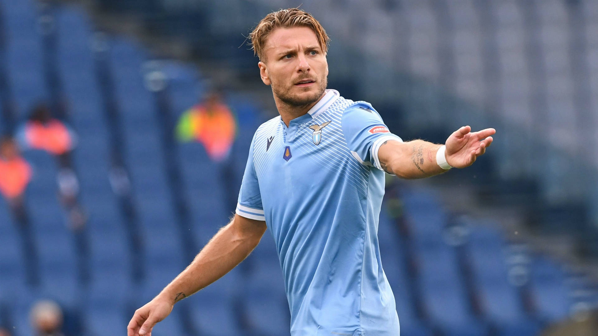 ciro immobile 桌布 壁紙 — 一個專注的足球運動員身穿淺藍色球衣，在身後空藍色座位的體育場上強烈地做手勢。