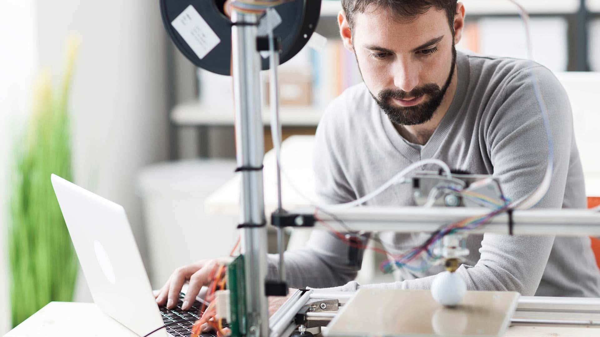 3d Printer 桌布 壁紙 — 一個專注的鬍鬚男穿著灰色襯衫，在3D印表機旁邊用筆記型電腦工作，印表機有彩色電線和一卷線材。