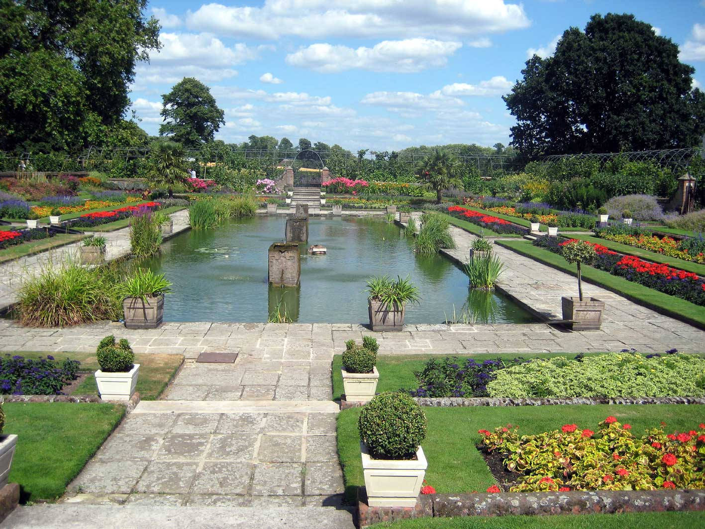 Garden 桌布 壁紙 — 一個對稱的正式花園，中央設有水景，兩旁是色彩繽紛的花壇和修剪整齊的樹籬，在明亮的藍天下。