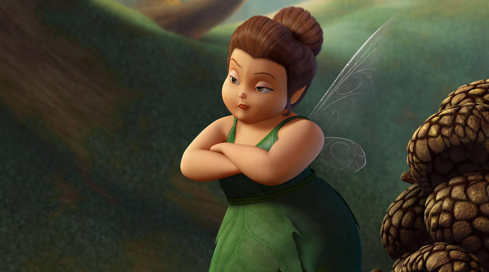 Tinker Bell 桌布 壁紙 — 一個小動畫仙子，棕色頭髮紮成髻，有透明翅膀，在森林裡松果附近交叉手臂站著。