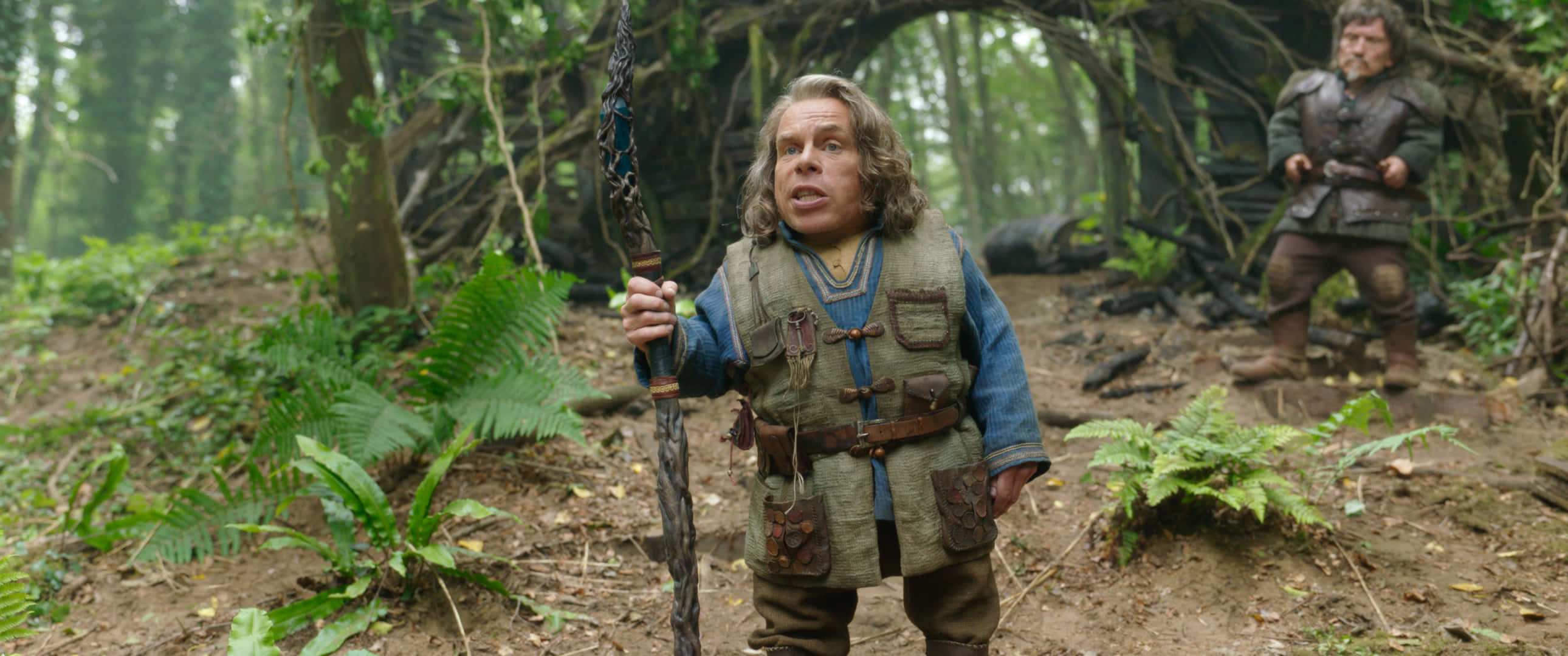 Warwick Davis 桌布 壁紙 — 一個小型奇幻戰士手持法杖在迷霧森林中警覺地站立，而另一個身影從纏結的樹根後注視著。