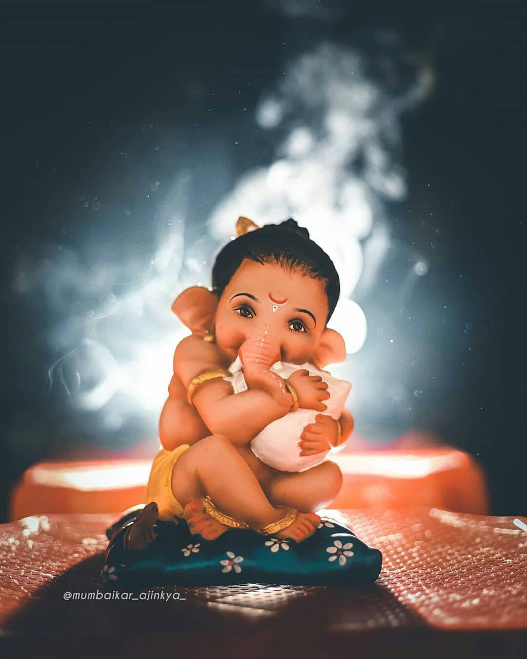 cute ganesha 桌布 壁紙 — 一個小型的象神雕像盤腿坐在花卉坐墊上，擁抱著一塊白布，背景煙霧繚繞、微微發光。