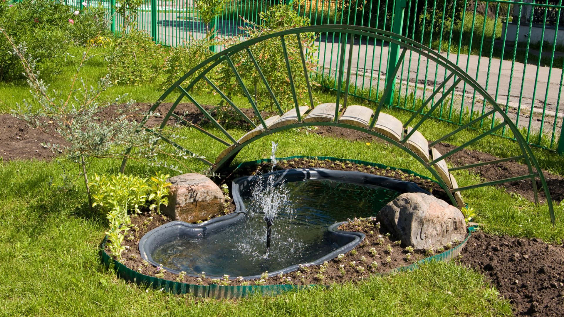 Garden Design 桌布 壁紙 — 一個小型花園噴泉，帶有彎曲的綠色金屬拱門和石頭裝飾，在草地後院噴水。