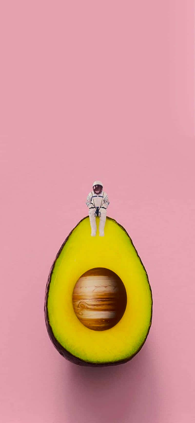 Avocado iPhone 桌布 壁紙 — 一個小太空人坐在半個酪梨上，其果核內可見木星，背景為柔和粉紅色。